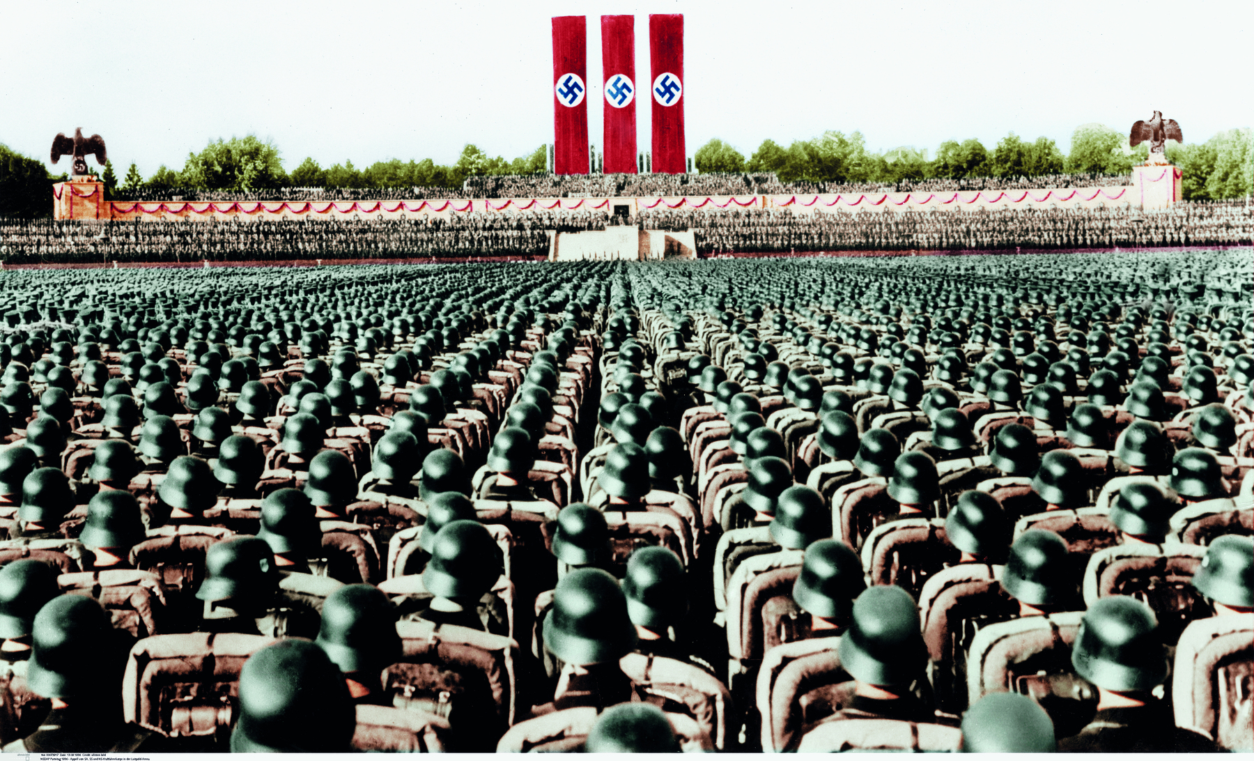 Fotografia. Inúmeras fileiras de soldados, com os corpos voltados em direção a um palanque, centralizado e ao fundo, contendo três faixas verticais idênticas, com fundos vermelhos e o símbolo da suástica.
