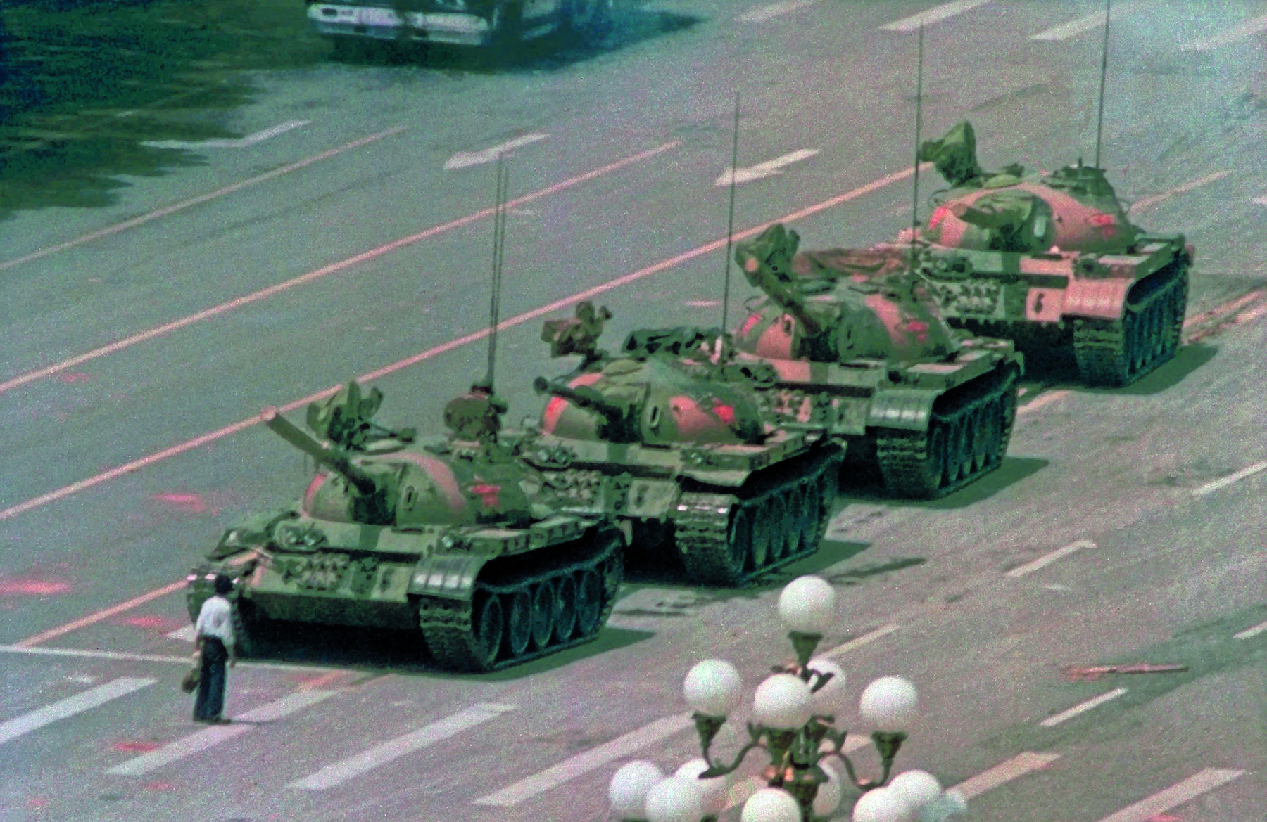 Fotografia. Destaque para uma pessoa em pé, vista de costas, sobre uma via asfaltada, à frente de quatro tanques de guerra com canhões, enfileirados.