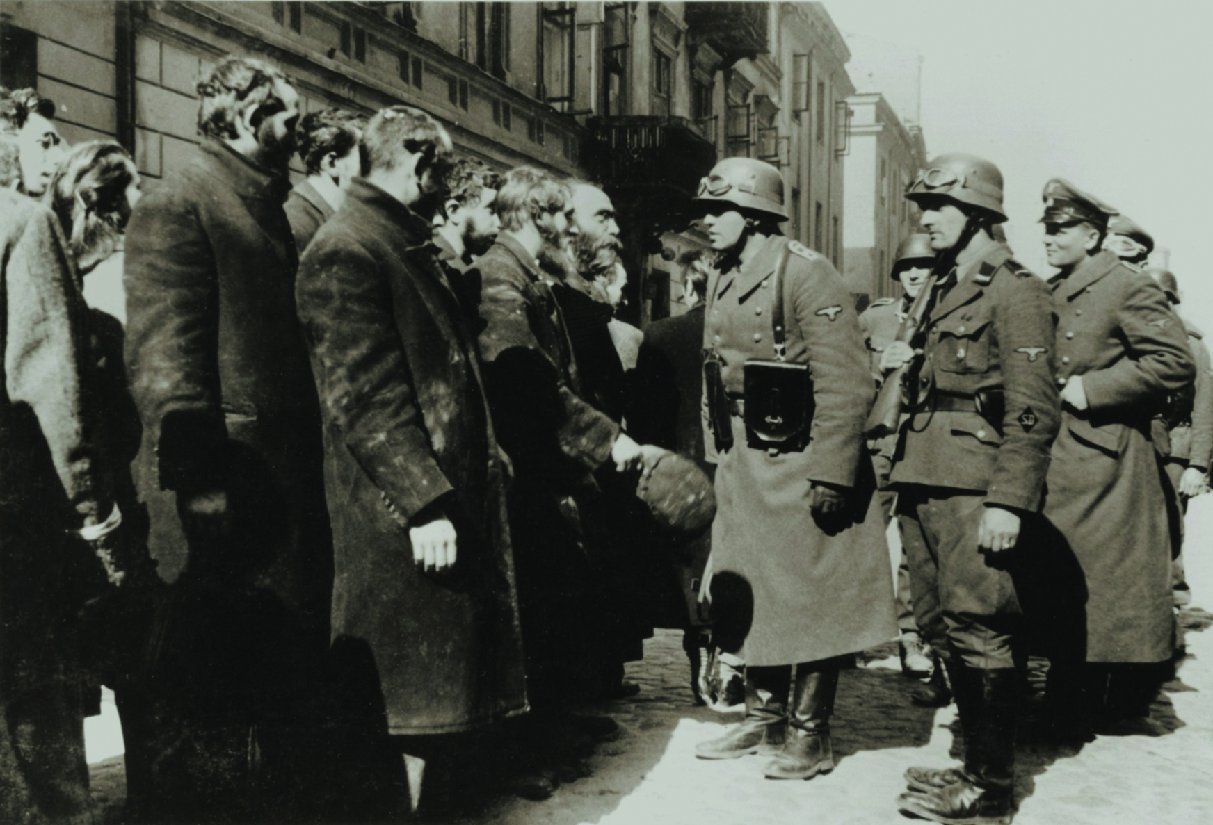 Fotografia em preto e branco. Sobre uma via, à direita, alguns homens vestindo fardas e capacetes. Na frente deles, à esquerda, um grupo de homens reunidos, vestindo casacos.