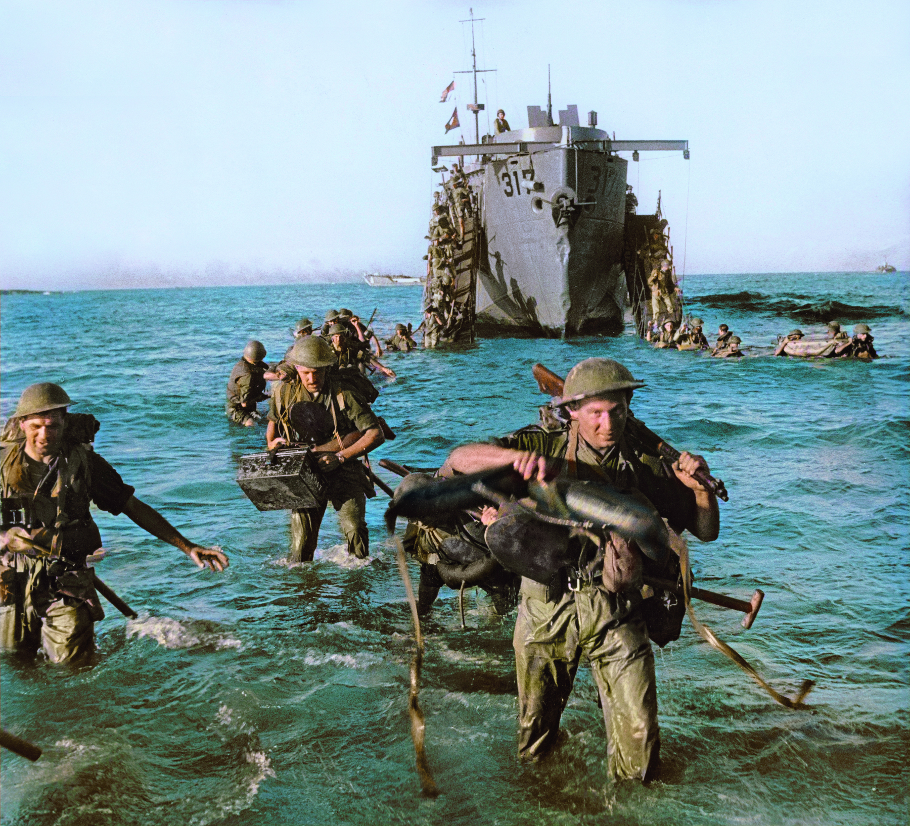 Fotografia. Soldados fardados, com água do mar até a altura dos joelhos, carregando armas, equipamentos e munição. Em segundo plano, sobre as águas, um navio de guerra de casco acinzentado.