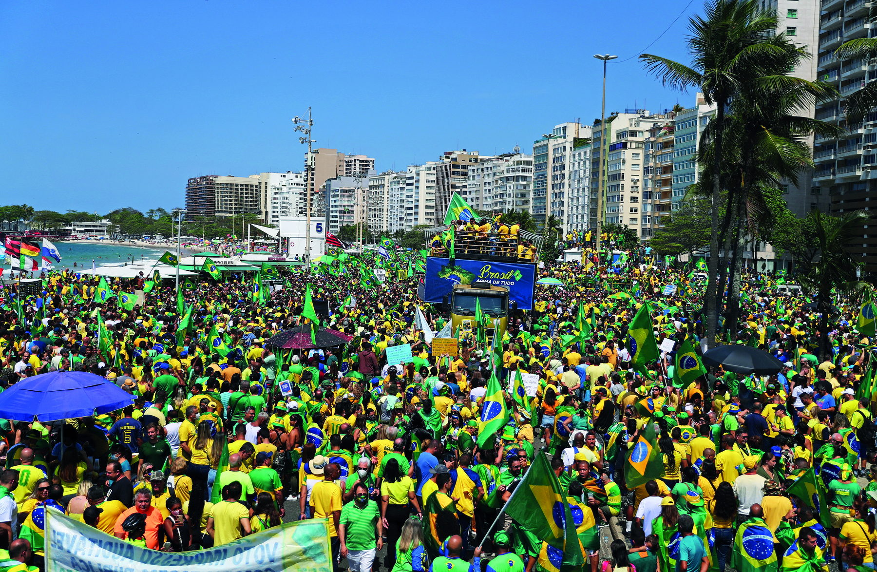 Fotografia. Em uma praia, rodeada por prédios altos, uma multidão de pessoas vestindo camisetas amarelas e verdes, portando bandeiras do Brasil. No centro da manifestação há um caminhão de trio elétrico, com pessoas sobre ele.  Ao fundo, no canto esquerdo, o mar.