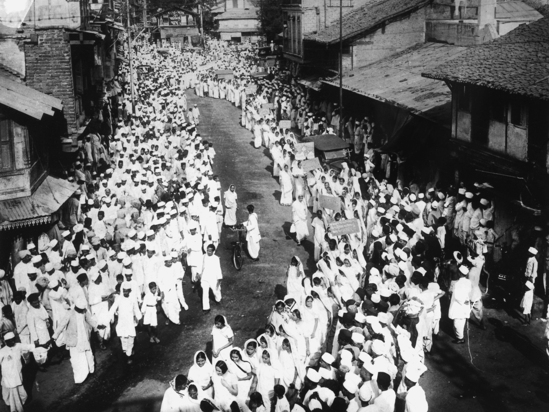 Fotografia em preto e branco. Vistas de cima, centenas de pessoas aglomeradas e dispostas em filas em uma rua, todas trajando vestimentas claras.