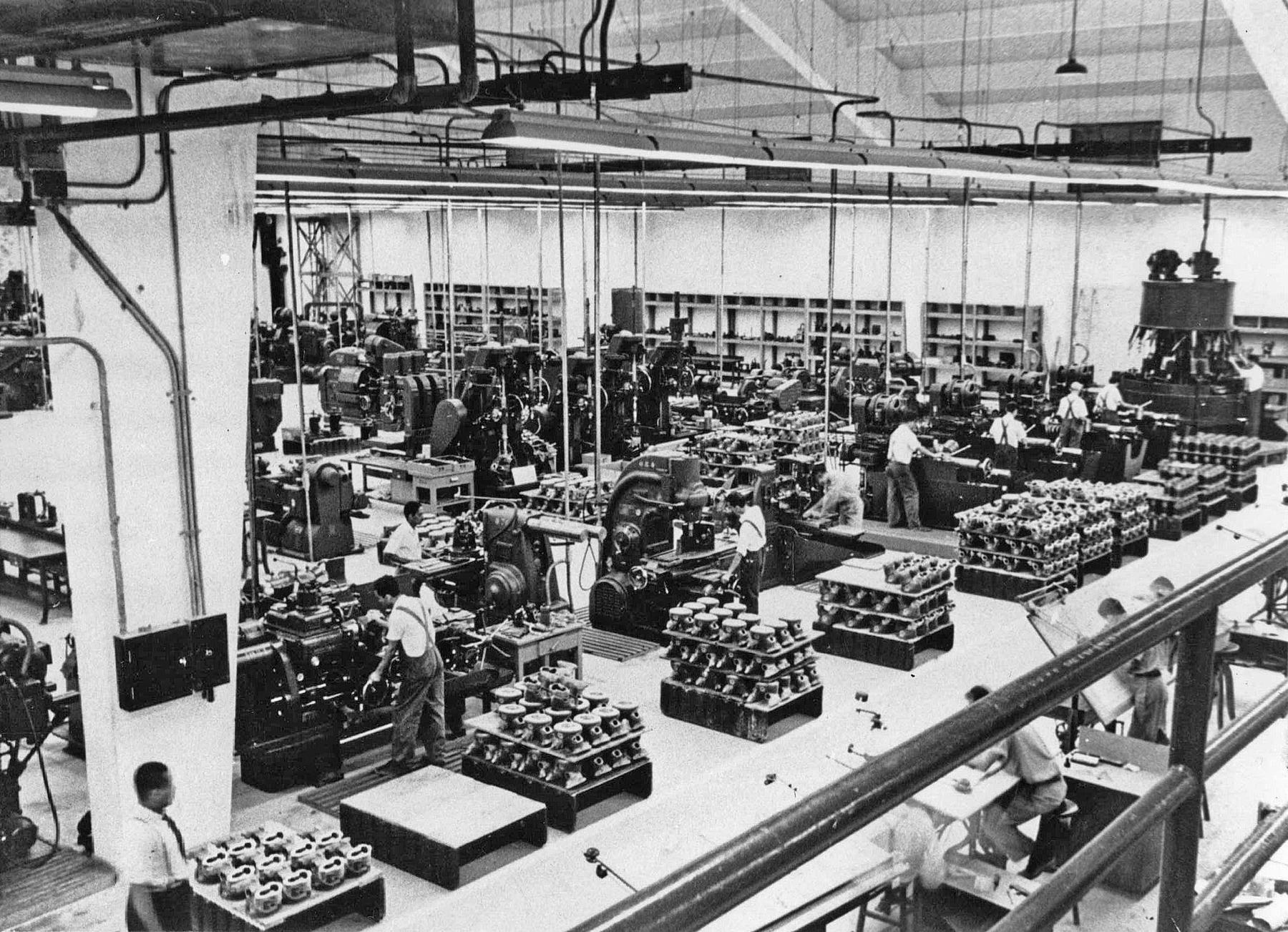 Fotografia em preto e branco. Vista do interior de uma fábrica, contendo maquinário, peças diversas, e homens por entre os corredores.