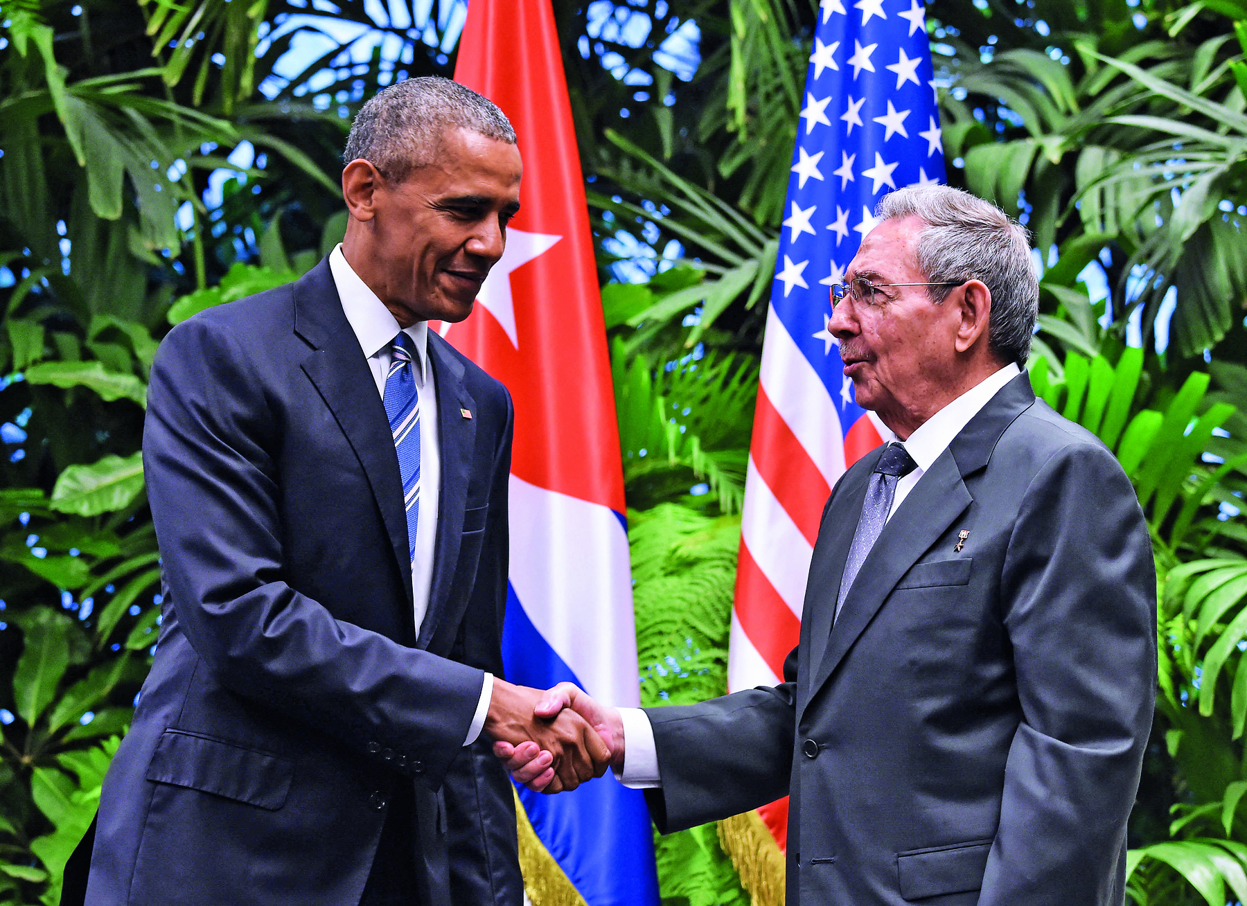Fotografia. Dois homens se cumprimentando com um aperto de mãos. O da esquerda, tem cabelos curtos e grisalhos, veste um terno escuro e uma gravata azul. O da direita, tem cabelos curtos, lisos e grisalhos, e veste um terno cinza. Em segundo plano, duas bandeiras, uma de Cuba e uma dos Estados Unidos.