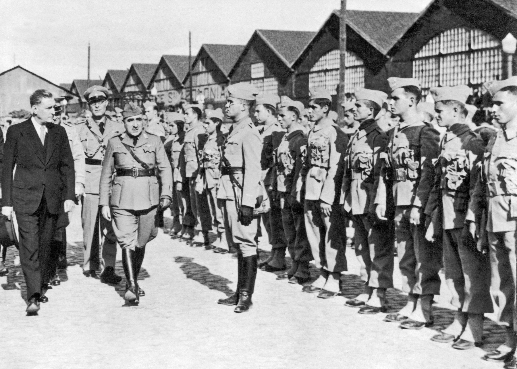 Fotografia em preto e branco. Destaque para um homem de terno escuro, à esquerda, acompanhado de alguns homens fardados, caminhando em frente a uma fileira de soldados. Ao fundo, galpões com tetos triangulares.