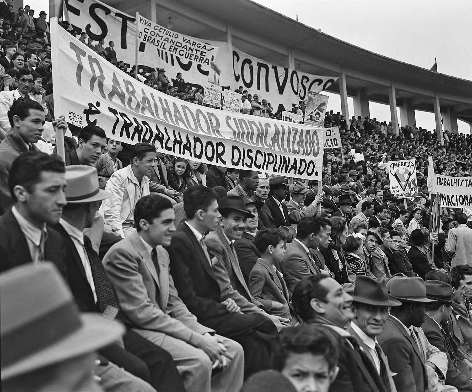Fotografia em preto e branco. Uma multidão de pessoas sentadas lado a lado e arquibancadas, portando faixas diversas.