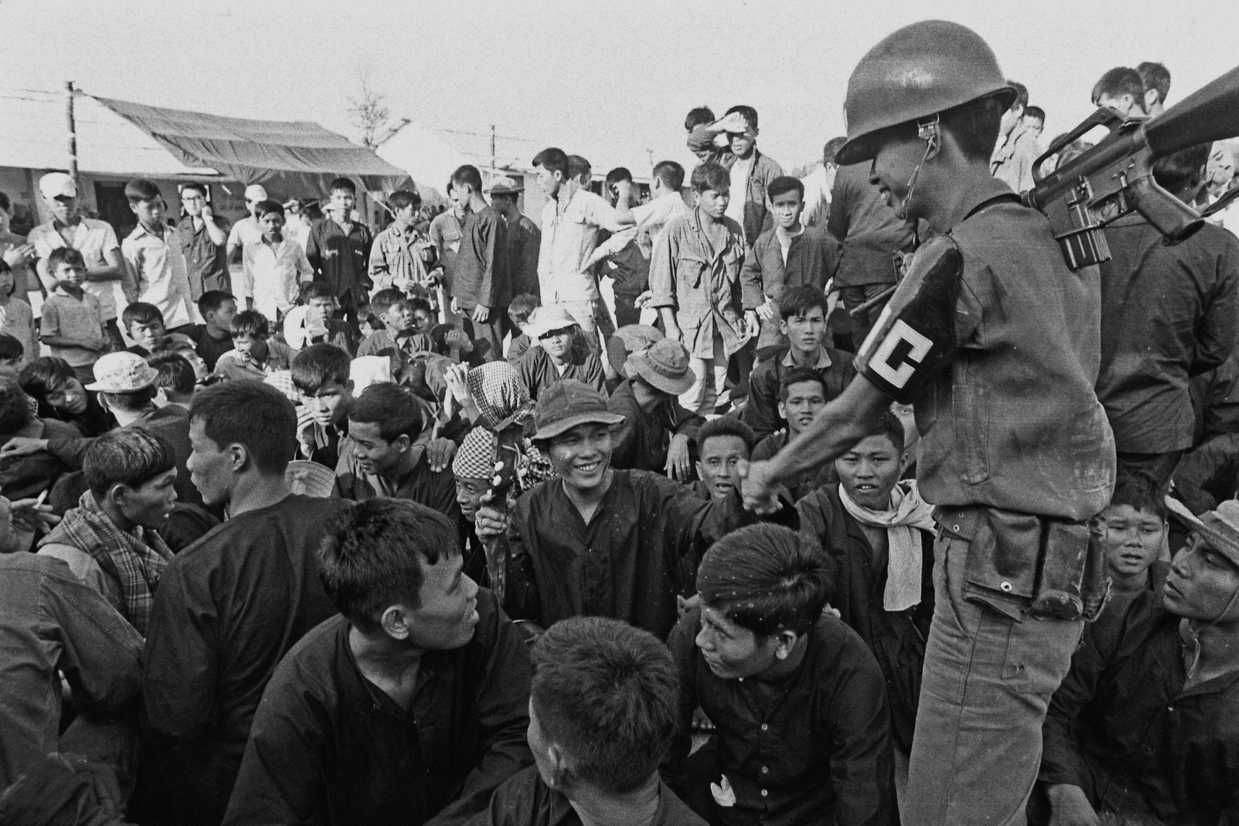 Fotografia em preto e branco. Diversos pessoas reunidas, próximas umas às outras. Na lateral direita da imagem, um soldado fardado aperta a mão de um homem.