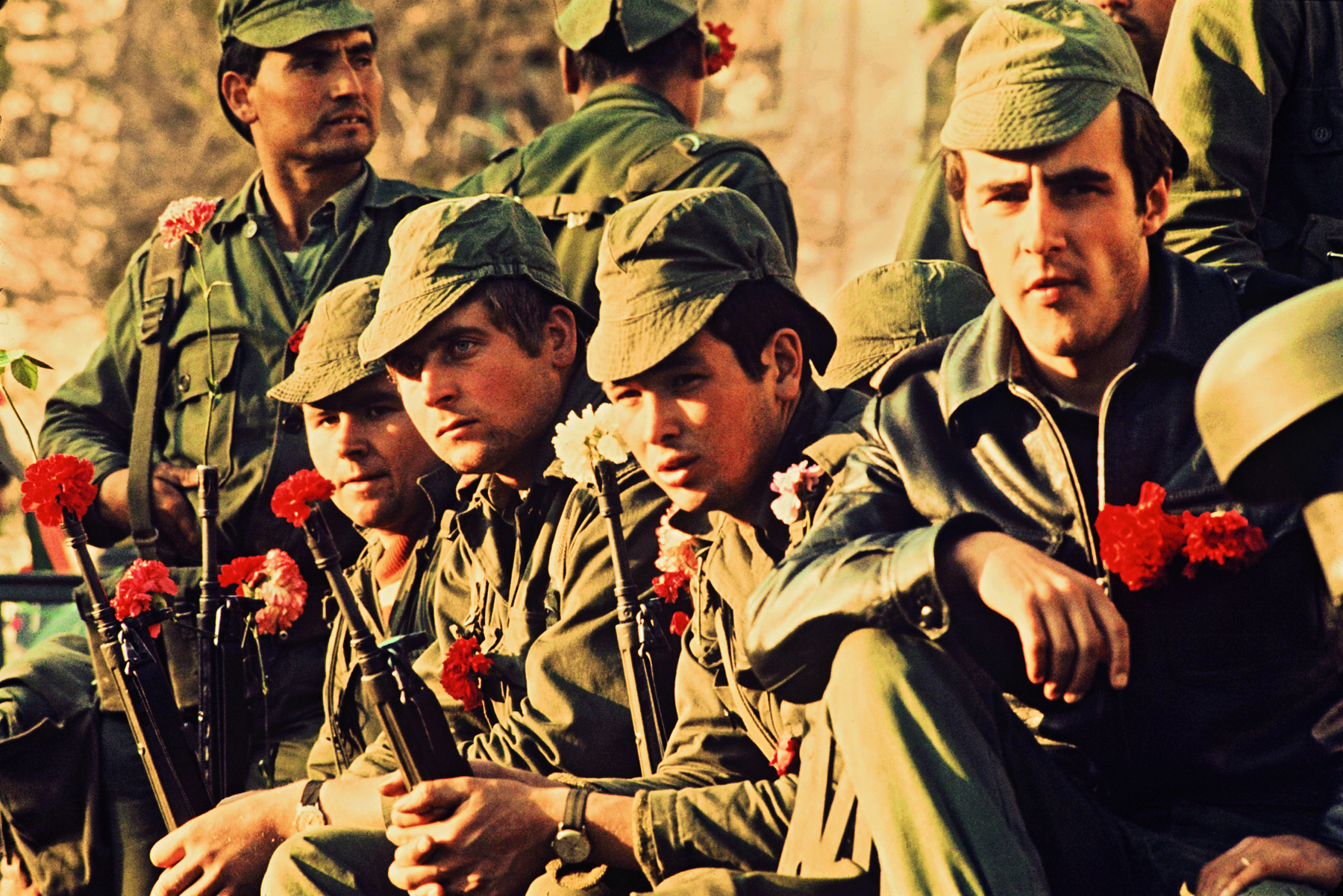 Fotografia. Soldados lado a lado, vestindo fardas e bonés, portando armas de fogo com flores vermelhas e brancas nas pontas.