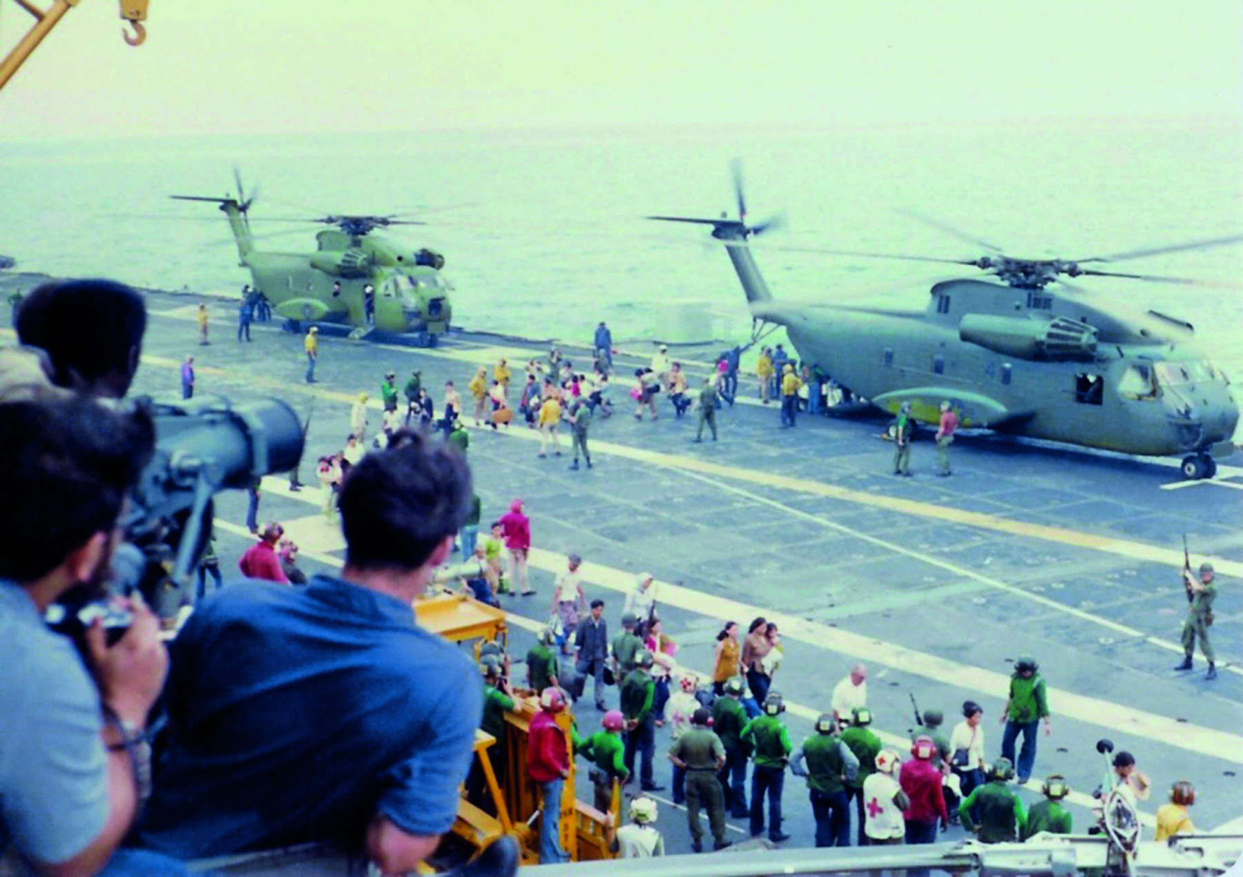 Fotografia. Dois helicópteros militares pousados na pista de um porta-aviões. Algumas pessoas caminham em direção às aeronaves enquanto um grupo de pessoas, no primeiro plano da imagem, observa a cena. Alguns portam máquinas fotográficas.