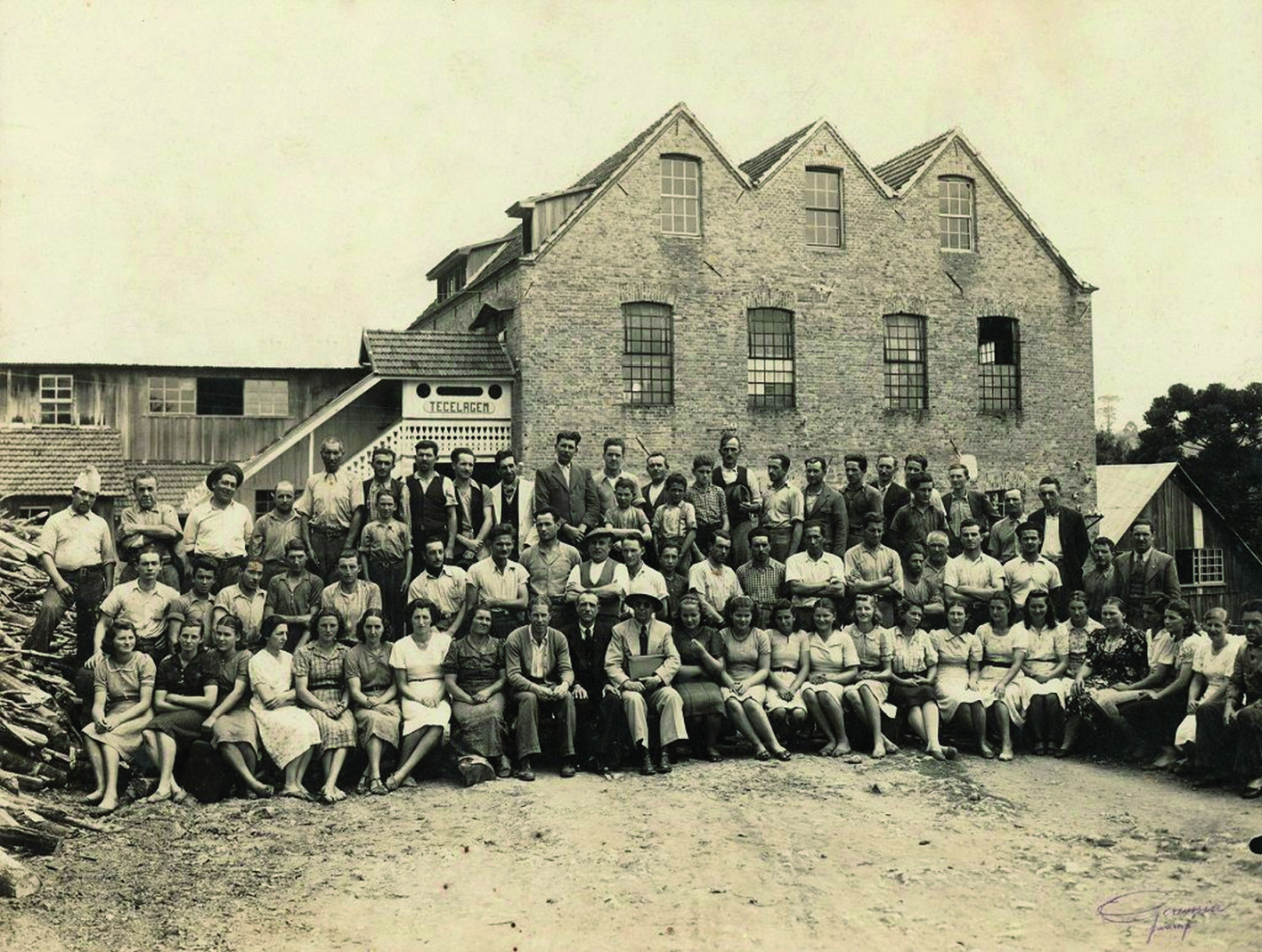 Fotografia em sépia. Em frente a fachada de uma fábrica, um grupo de pessoas reunidas, enfileiradas lado a lado. À frente, sentadas. E atrás outras duas filas de pessoas em pé.