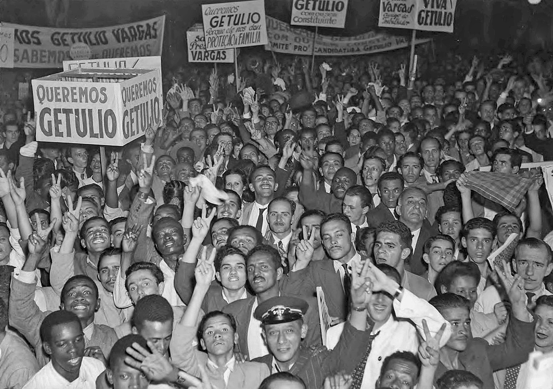 Fotografia em preto e branco. Uma multidão de pessoas aglomeradas, portando faixas e cartazes com o texto: 'Queremos Getúlio'.