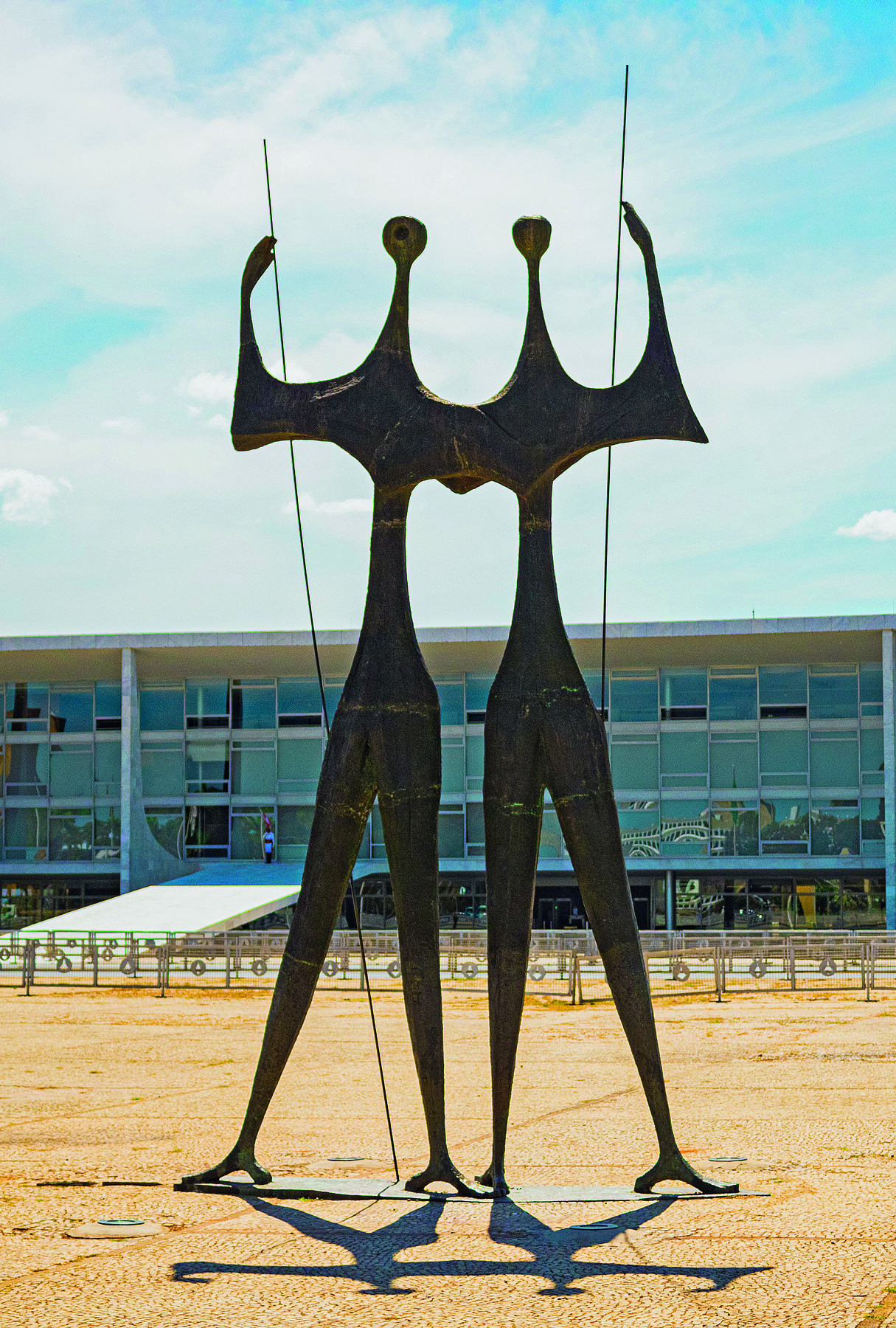 Fotografia. Em uma praça, diante de uma construção de janelas envidraçadas, uma escultura retratando dois personagens de aspectos humanos, em pé, com os braços dados, portando uma haste cada um.