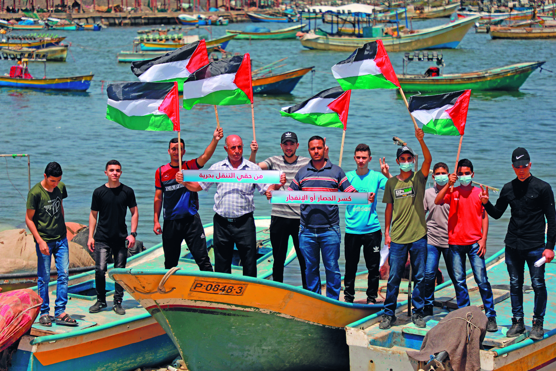 Fotografia. Um grupo de homens, vistos de frente, lado a lado, sobre algumas pequenas embarcações, portando bandeiras da Palestina. A bandeira da Palestina é retangular, possui um triângulo vermelho à direita e três faixas, uma preta, uma branca e uma verde, de cima para baixo. 