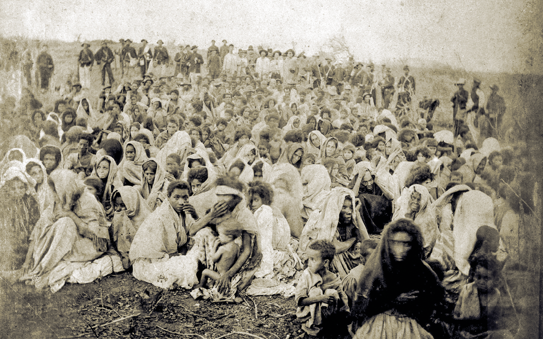 Fotografia em preto e branco. Uma multidão de pessoas aglomeradas, sentados no solo, lado a lado, a maioria vestindo roupas claras, e ao fundo, um grupo de homens em pé, armados, cercando o perímetro.