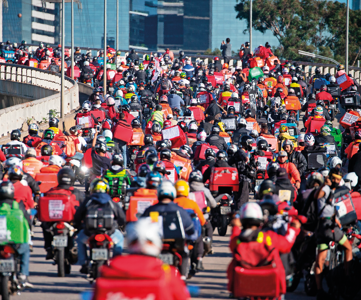 Fotografia. Uma multidão de pessoas aglomeradas sobre uma via pavimentada, todos sobre motocicletas, a maioria utilizando capacetes e portando mochilas quadradas sobre as costas ou baús sobre as motocicletas.