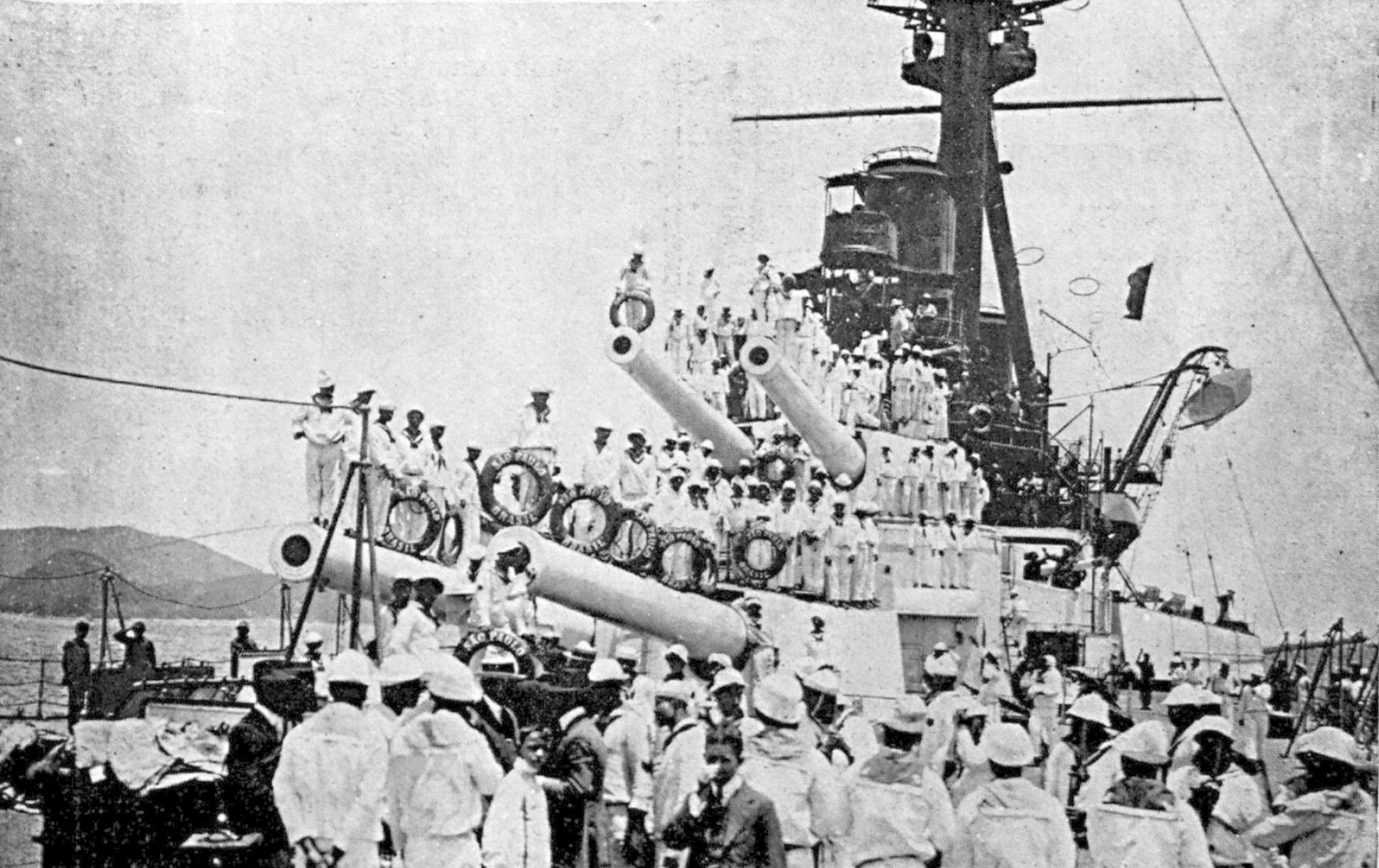 Fotografia em preto e branco. Um navio de guerra com diversos canhões, repleto de homens vestindo calças, camisas e boinas brancas, com lenços ao redor do pescoço.