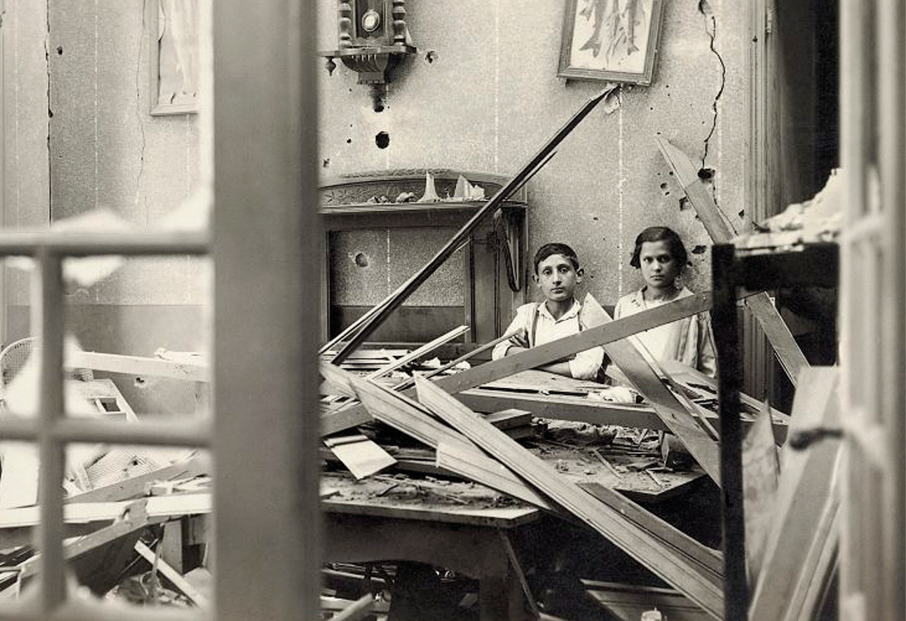 Fotografia em preto e branco. Duas pessoas, sentadas lado a lado, no interior de um cômodo em destroços, com paredes rachadas, pedaços de madeira sobre o chão, e móveis destruídos.
