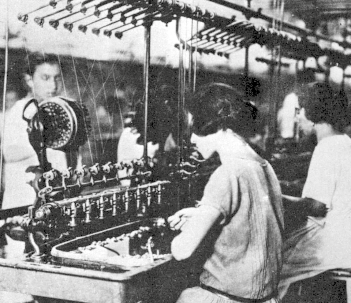 Fotografia em preto e branco. Duas mulheres, lado a lado, sentadas a frente de uma máquina de tecelagem, com fios suspensos e hastes metálicas.