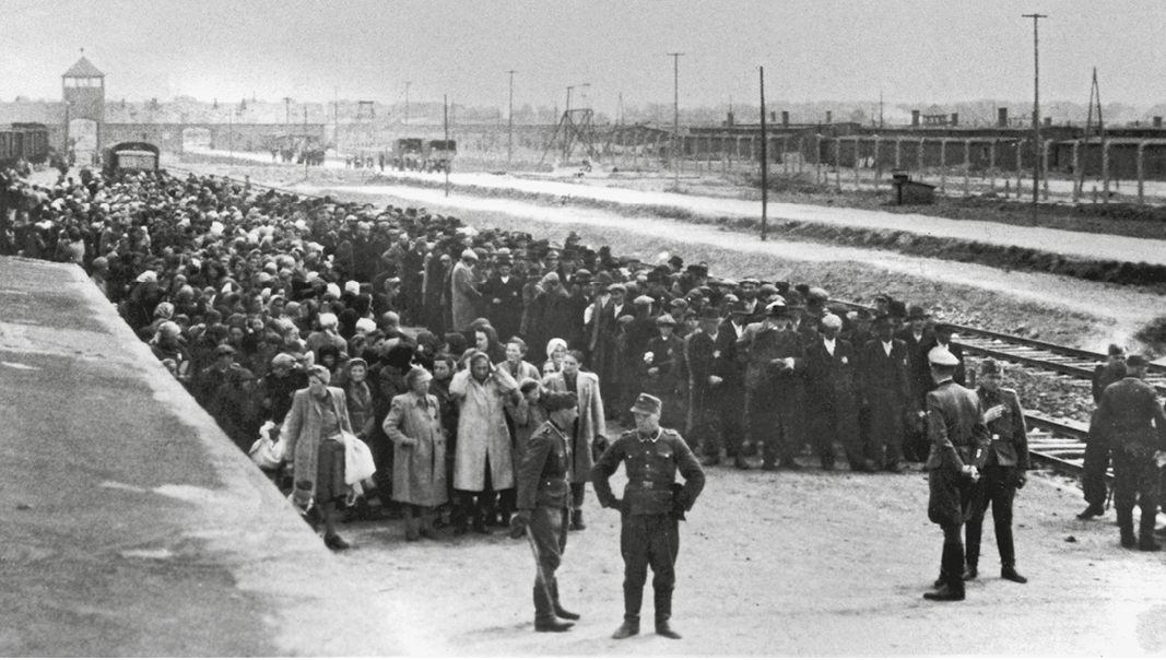 Fotografia em preto e branco. Uma multidão de pessoas aglomeradas sobre uma passarela. À frente da multidão, homens fardados.