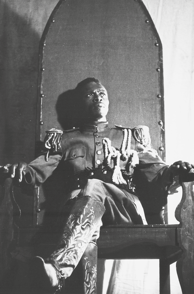 Fotografia em preto e branco. Um homem visto de frente, com cabelos curtos e escuros, vestindo uma farda e uma calça, sentado sobre um trono, apoiando suas mãos nos encostos laterais, e com as pernas cruzadas.