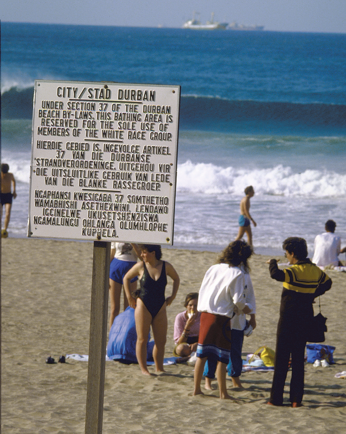 Fotografia. Praia com faixa de areia clara, e o mar azul ao fundo, destaque para uma placa contendo textos. Sobre a faixa de areia, algumas pessoas em pé e outras sentadas sobre toalhas.