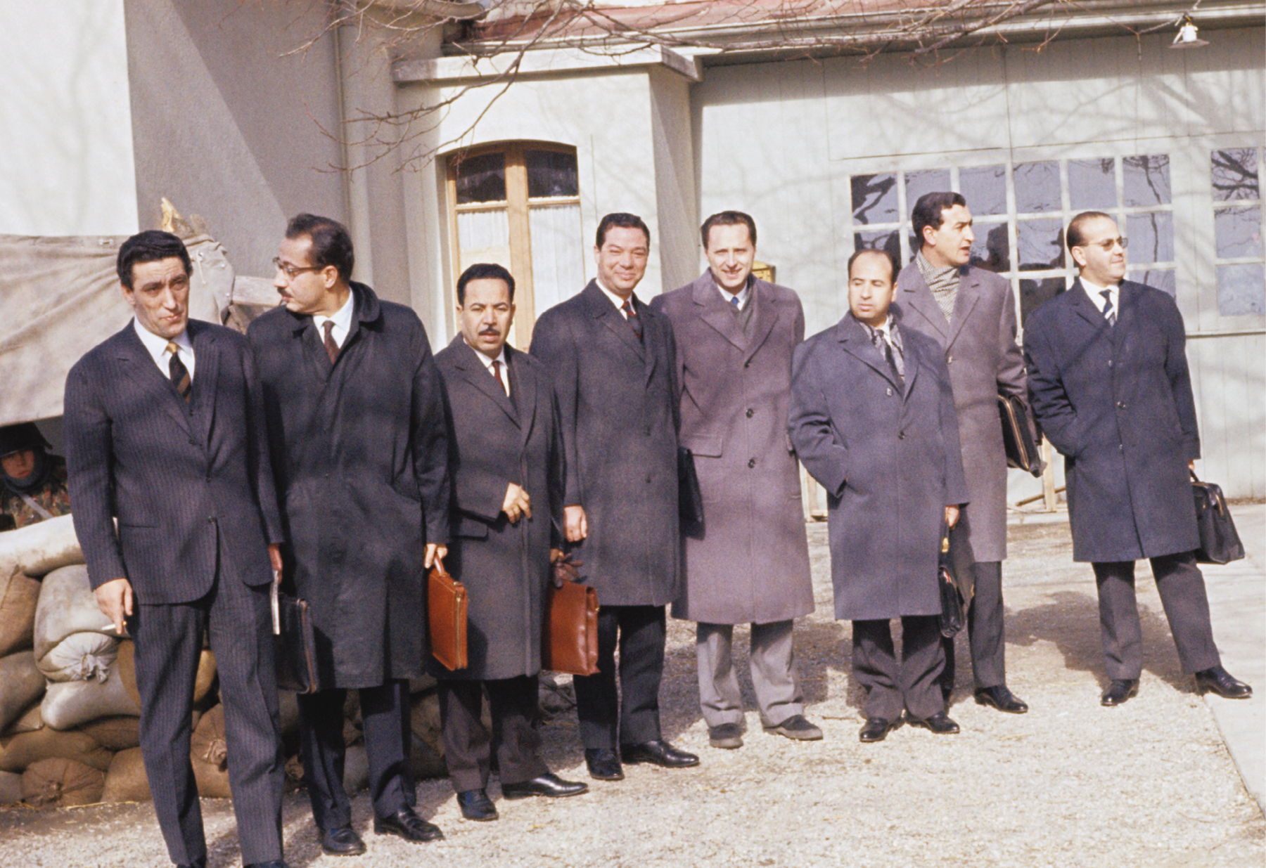 Fotografia. Um grupo de homens, em pé, lado a lado, todos vestindo paletós ou casacos escuros. Em segundo plano, a fachada de uma casa vista parcialmente, contendo entre as paredes janelas e vitrais quadrados.