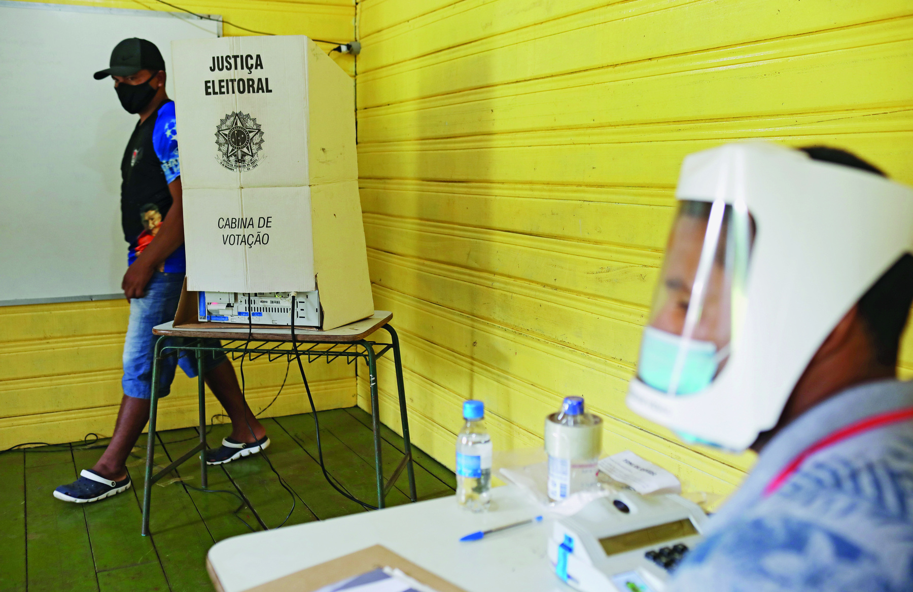 Fotografia. Em uma sala fechada, de parede amarelo clara e piso verde, ao fundo, há um homem em pé, que veste uma camiseta preta e azul e bermuda jeans. Ele usa um boné preto e uma máscara preta cobrindo a boca e o nariz. 
O homem está com o corpo voltado para a esquerda, próximo a uma cabine de votação disposta sobre uma carteira escolar. A cabine é branca e, nela estão gravados  os textos: 'Justiça eleitoral', na parte superior, o brasão da república no centro, e  'Cabina de votação' na parte inferior. 
A cabine tem uma abertura inferior, que mostra a parte posterior de uma urna eletrônica e sua fiação.
À frente, com um efeito desfocado, há um homem sentado diante de uma mesa. Ele usa uma camiseta cinza, uma máscara cirúrgica azul sobre o nariz e a boca e um 'face shield' branco a frente de seu rosto. 
Sobre a mesa, uma prancheta, uma caneta, uma garrafa de água e um aparelho eletrônico com um teclado numérico.