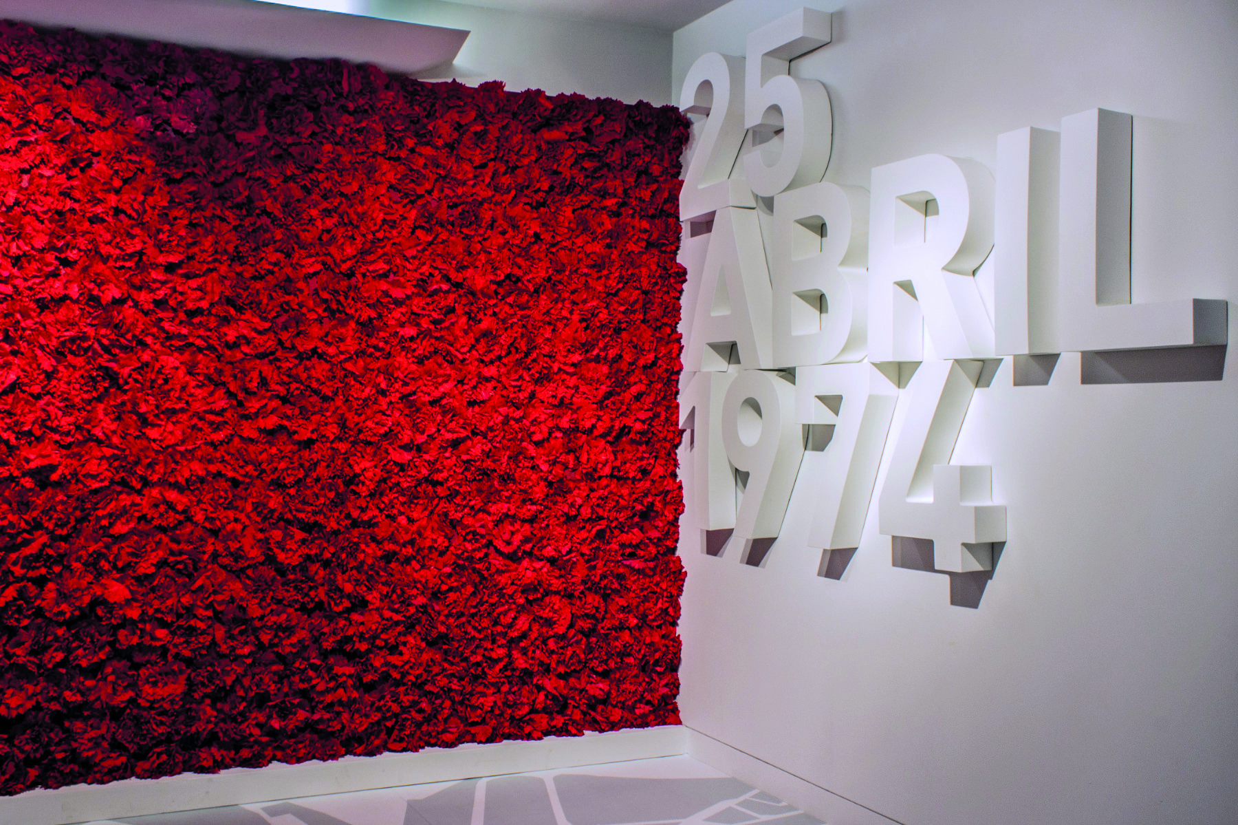 Fotografia. Um mural de superfície vermelha e textura simulando flores. Ao lado, o texto: '25 ABRIL 1974', escritos em relevo sobre a parede.