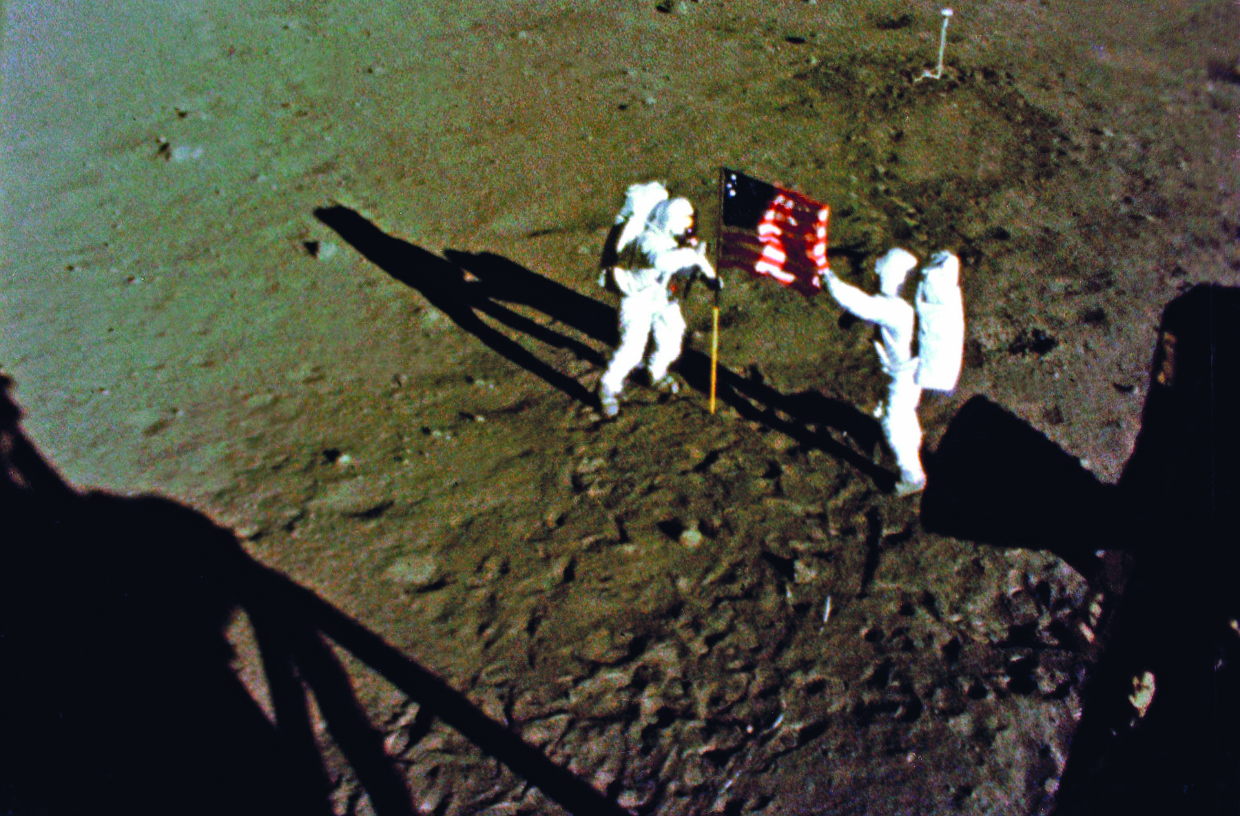 Fotografia. Duas pessoas vestindo trajes de astronautas, de coloração branca, cobrindo todas as partes de seus corpos e capacetes. Estão em pé, sobre um solo terroso e acinzentado, e entre eles há uma haste vertical com uma bandeira dos Estados Unidos da América.