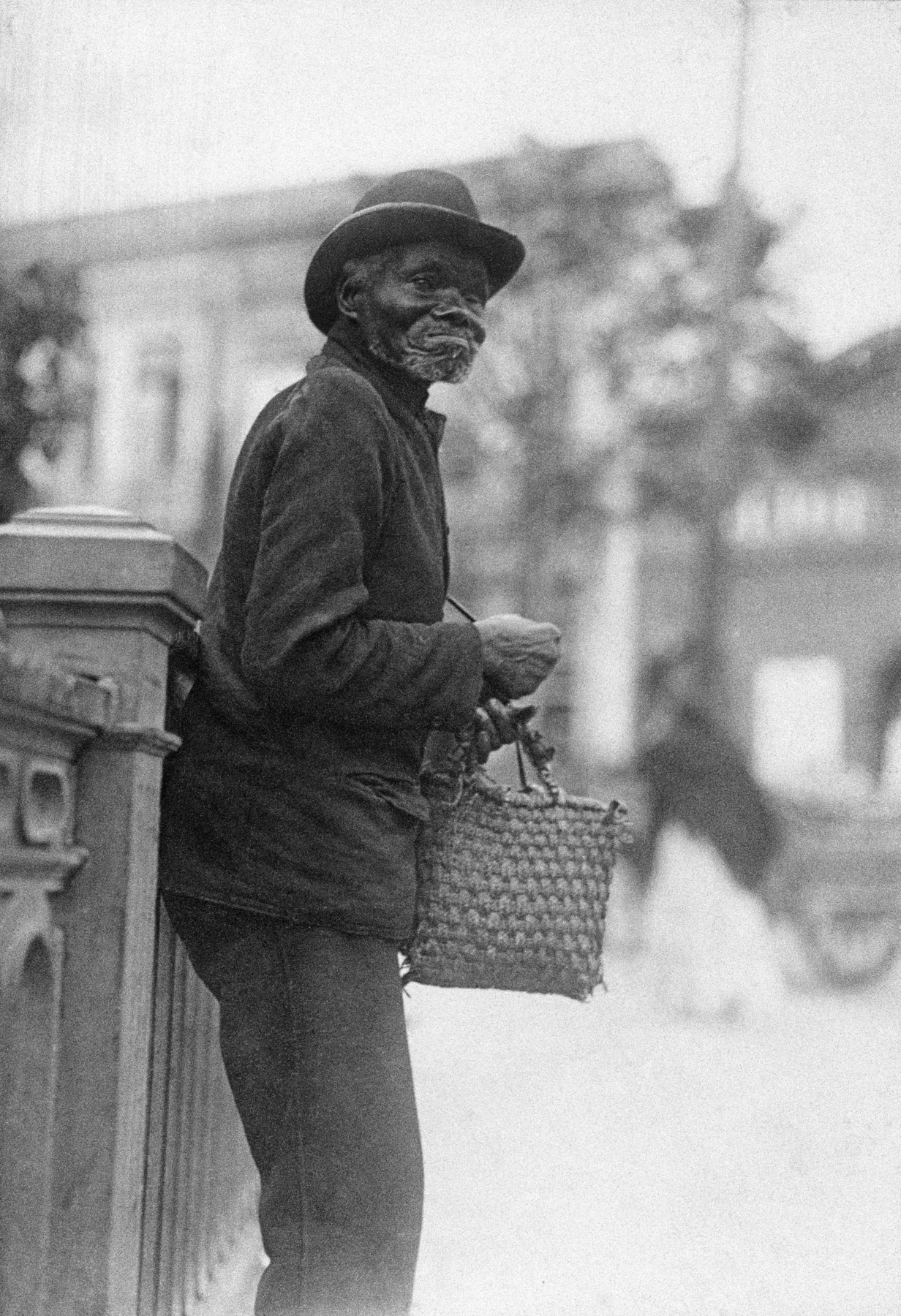 Fotografia em preto e branco. Destaque para um homem negro, idoso, em pé, visto de lado, vestindo uma blusa, uma calça e um chapéu com abas sobre sua cabeça. Segura uma pequena bolsa em uma de suas mãos.