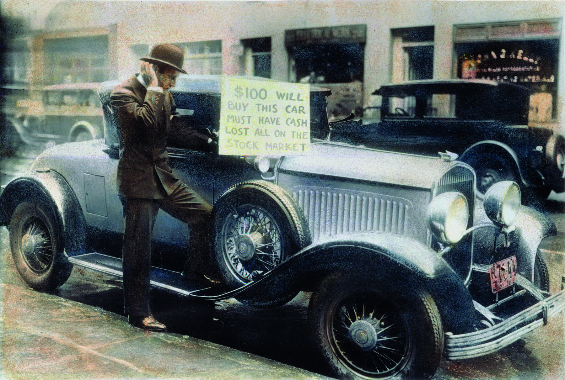 Fotografia. Um homem em pé, vestindo um paletó e calça marrons, e um chapéu com abas sobre a cabeça, ao lado de um automóvel de carroceria azulada, e uma placa branca contendo um texto.