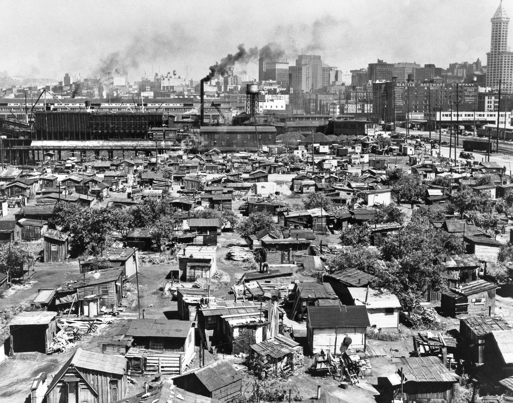 Fotografia em preto e branco. Vista aérea de uma zona urbana com diversos casebres lado a lado, amontoados sobre um grande terreno. Em segundo plano, fábricas e usinas, com chaminés expelindo fumaça.