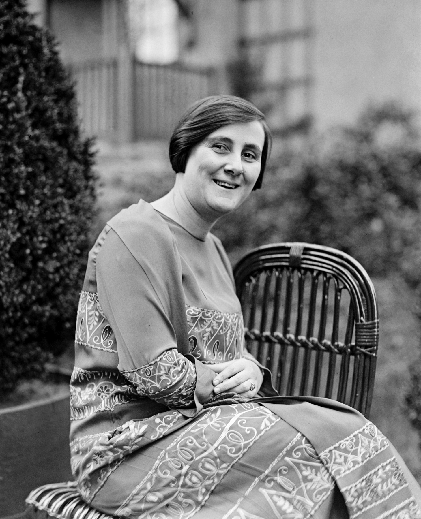 Fotografia em preto e branco. Uma mulher sorridente, de cabelos curtos e lisos, trajando um longo vestido, sentada de lado sobre uma cadeira.