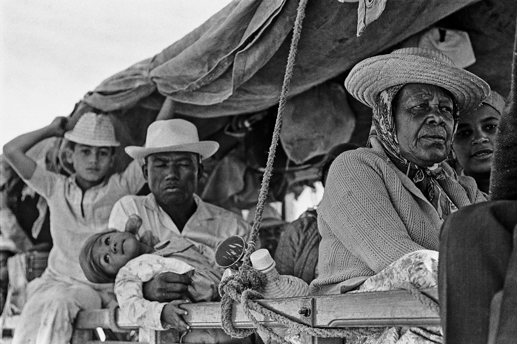 Fotografia em preto e branco. Um grupo de pessoas, entre homens, mulheres e crianças, sobre uma carroceira coberta com lonas de tecido. Vestem camisas e chapéus com abas.