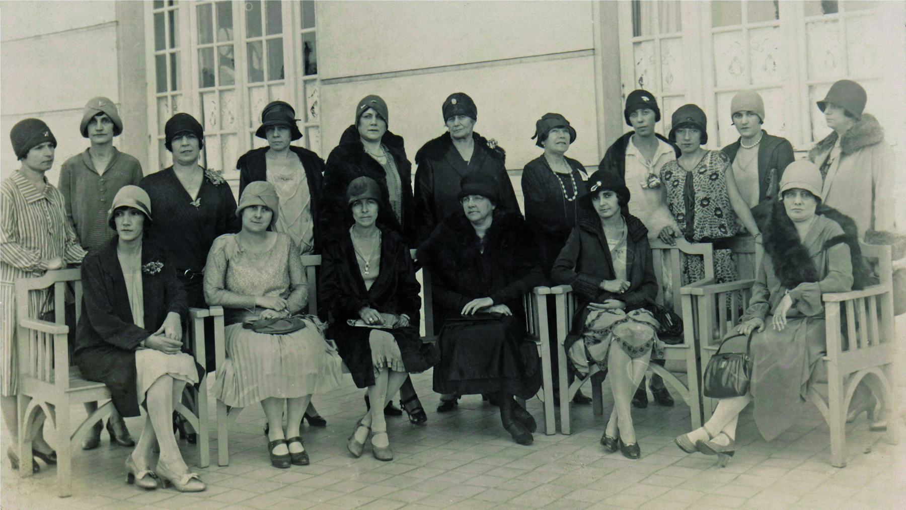 Fotografia em preto e branco. Um grupo de mulheres, lado a lado, distribuídas em duas fileiras. Na fileira da frente, as mulheres estão sentadas sobre cadeiras. Na fileira de trás, elas estão em pé. Todas elegantemente vestidas, trajando vestidos longos e chapéus. Algumas também usam casacos.