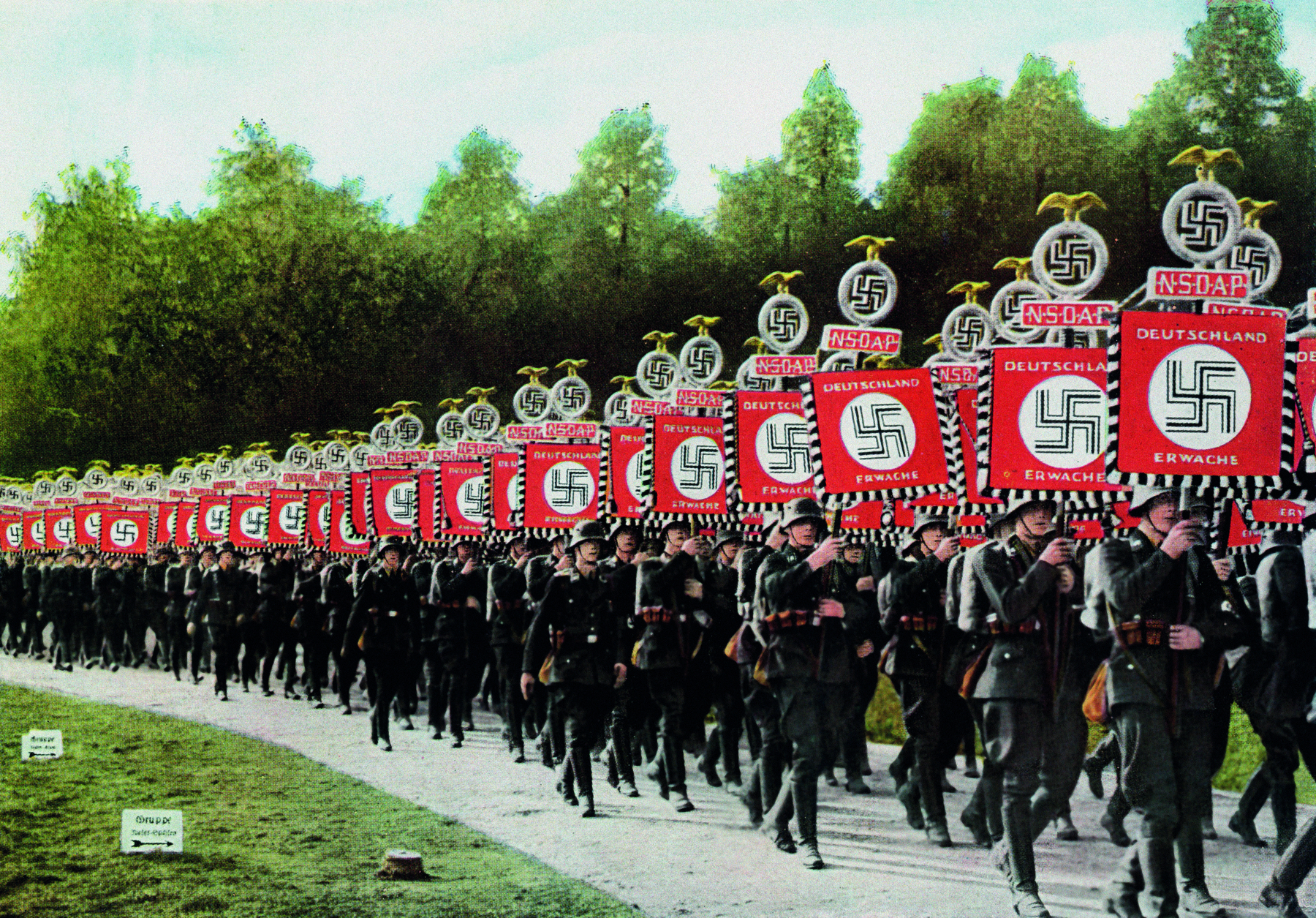 Fotografia. Soldados enfileirados, portando hastes com cartazes vermelhos com textos e símbolos de uma suástica.