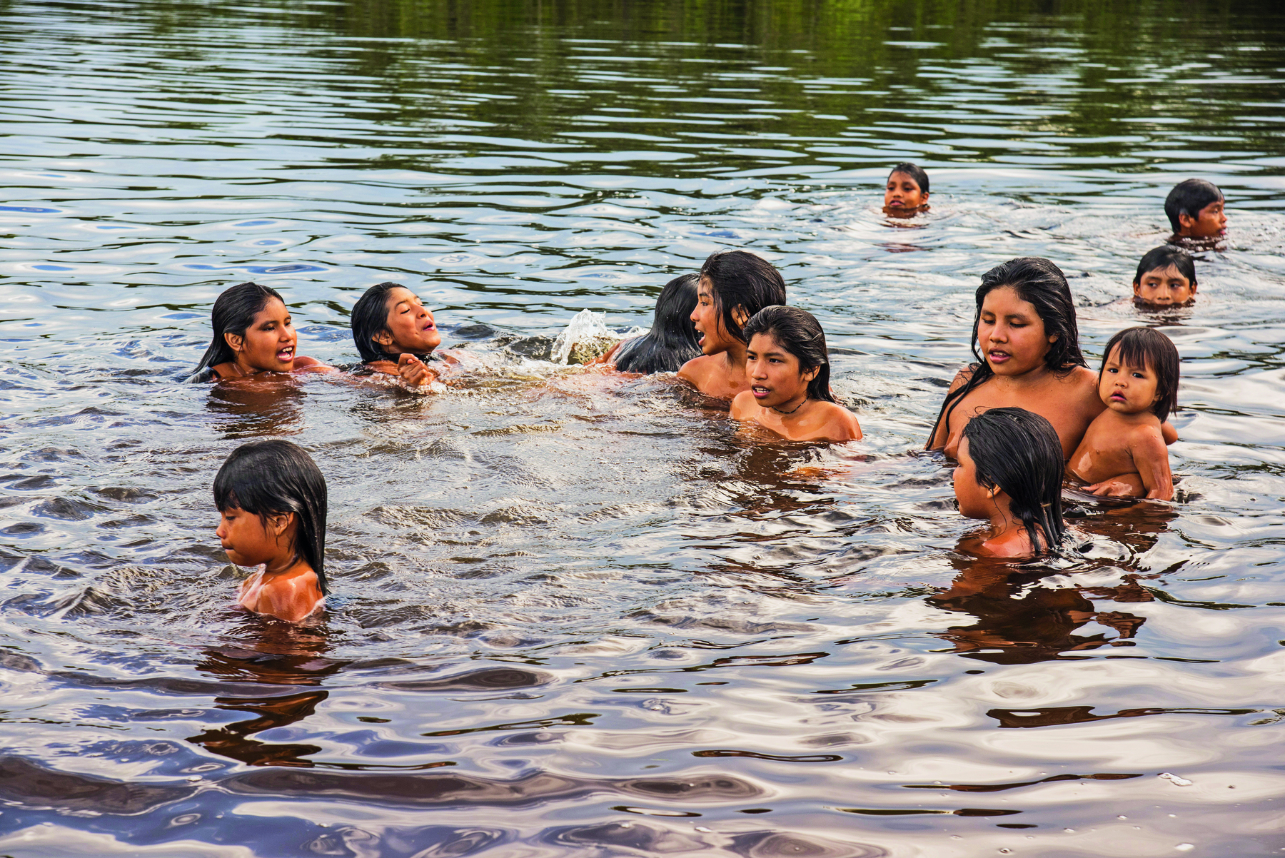 Fotografia. Crianças indígenas parcialmente submersas em um rio de águas levemente amarronzadas.