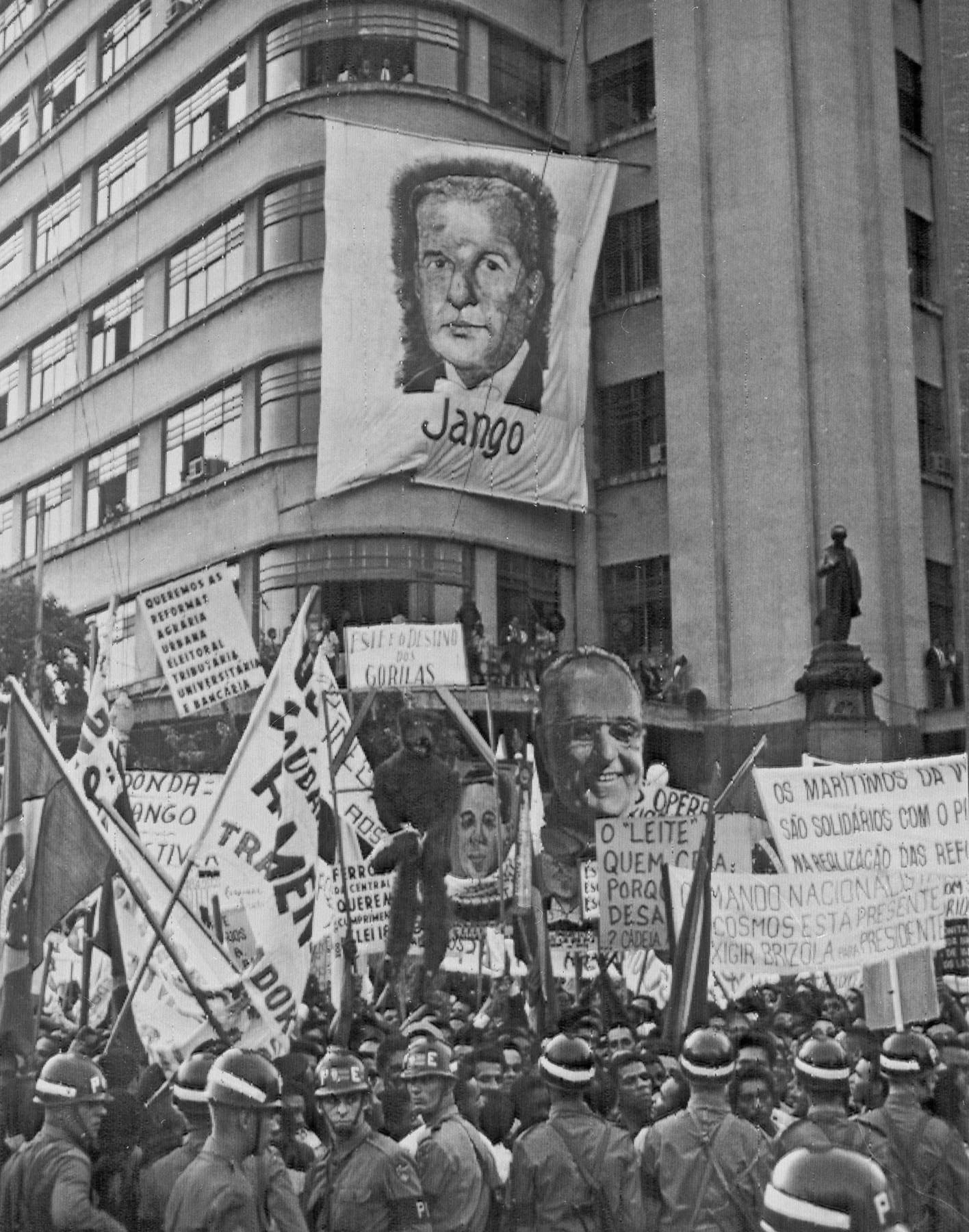 Fotografia em preto e branco. Uma multidão de pessoas aglomeradas em frente a um edifício, portando faixas e cartazes. Destaque para uma faixa, pendurada na fachada do edifício, retratando o rosto de um homem, com o nome: 'Jango'.