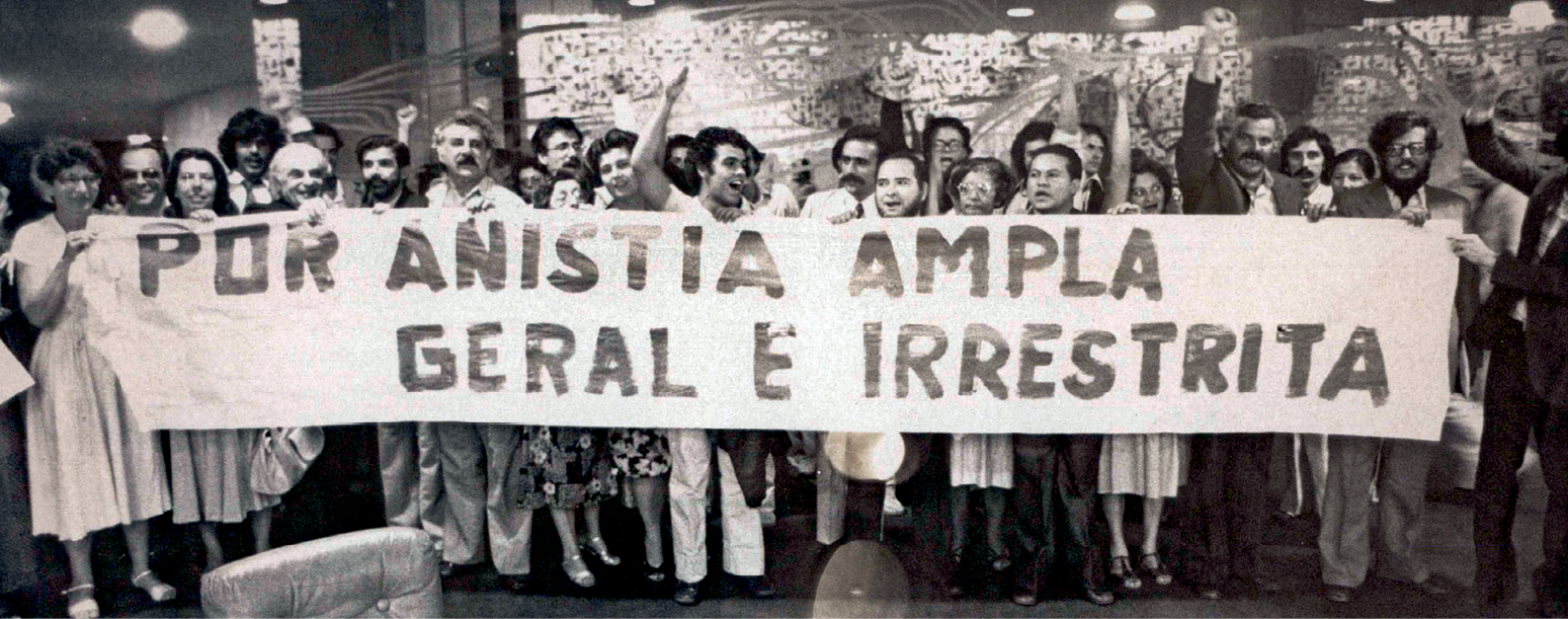 Fotografia em preto e branco. Em um espaço fechado, um grupo de pessoas, homens e mulheres, portando uma faixa com o texto: 'Por anistia ampla geral e irrestrita'.