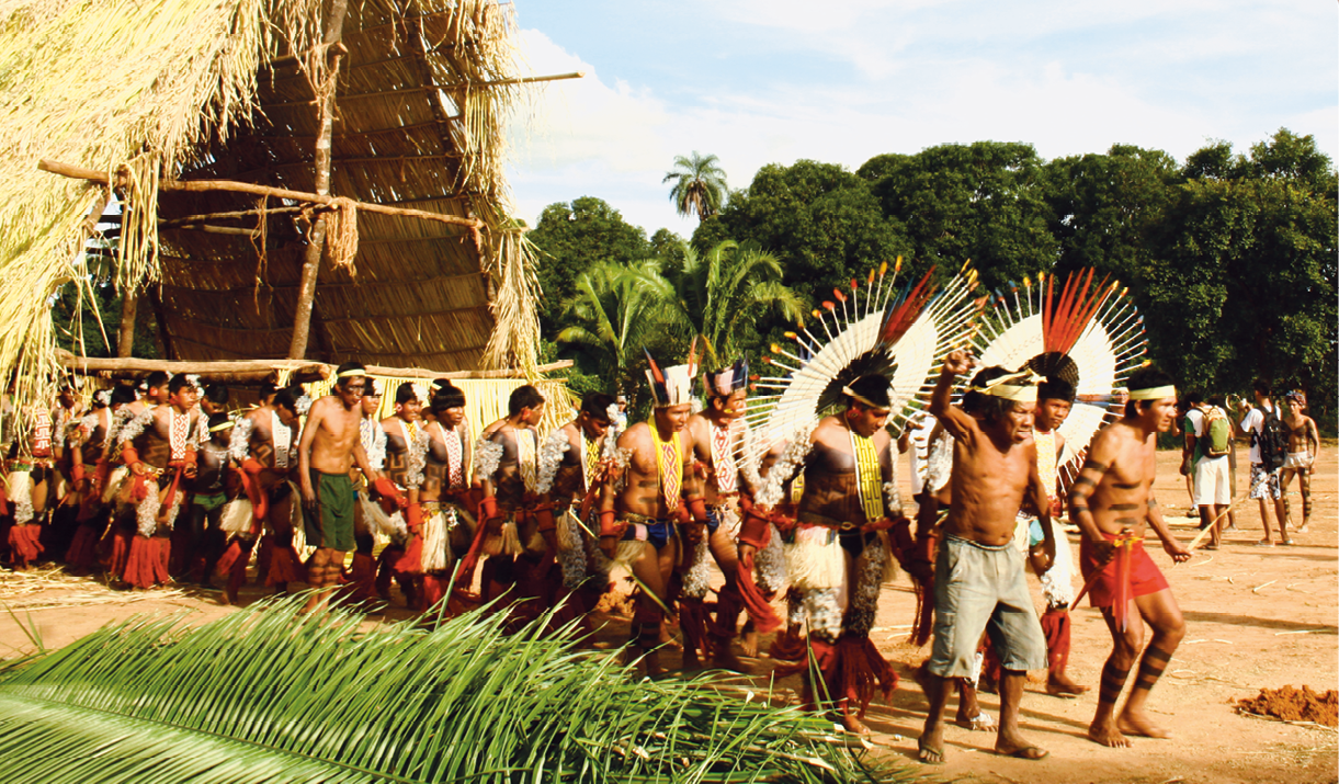 Fotografia. Sob um céu azul com nuvens, em um espaço aberto, com chão de terra e árvores ao fundo, um grupo de pessoas indígenas, majoritariamente homens de cabelos curtos, lisos e pretos, estão dispostos dois a dois em uma fila. Os homens à frente da fila usam bermudas, estão sem camisa, e têm faixas de cor clara amarradas em torno da cabeça. Os dois homens atrás deles, usam grandes adereços em formato circular feitos de penas brancas, com a parte central feita de penas vermelhas, e detalhes em amarelo nas pontas ao redor de todo o círculo. Eles também usam, preso ao pescoço, longos adereços coloridos com padrões geométricos; e outros adereços feitos de penas vermelhas amarrados em suas canelas. Atrás deles, há dois homens que usam cocares de penas coloridas e longos adereços coloridos com padrões geométricos no pescoço. Um deles, segura uma lança. Os demais homens em fila não usam cocares. A maior parte deles usa adereços longos, coloridos e com padrões geométricos presos ao pescoço, cobrindo a parte frontal do centro do corpo, além de braceletes na cor vermelha. Atrás deles, uma oca alta, feita de folhas e toras de madeira. Ao lado do grupo de homens, uma pilha de folhas verdes de palmeira.