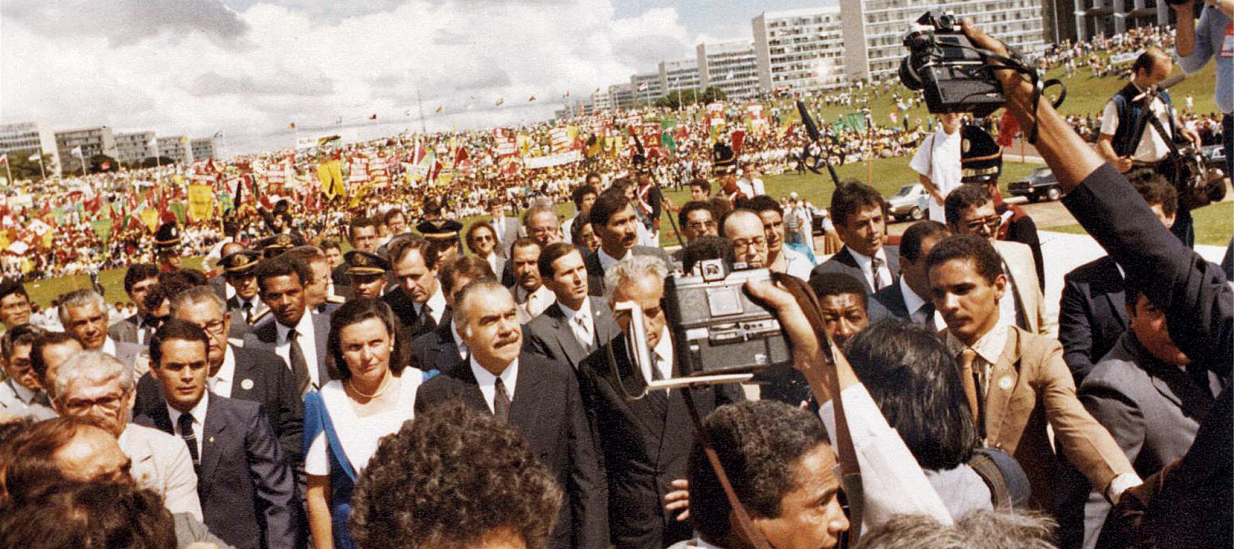 Fotografia. Em uma área aberta, gramada, ladeada por edifícios retangulares idênticos, ao fundo, uma multidão de pessoas em manifestação, carregando faixas e bandeiras. 
Há outro grupo, mais à frente, separado por homens com uniformes militares. Nesse grupo, em destaque, no centro, um homem, com cabelos curtos e grisalhos, um bigode escuro no rosto, vestindo terno escuro, camisa branca e gravata escura. À esquerda dele, uma mulher de cabelos médios, castanhos, lisos, usando um vestido branco com detalhes azuis, um colar e um par de brincos. Ao redor deles, dezenas de homens de terno e gravata. À direita da imagem, vistas de costas, duas pessoas com os braços levantados, segurando câmeras fotográficas.