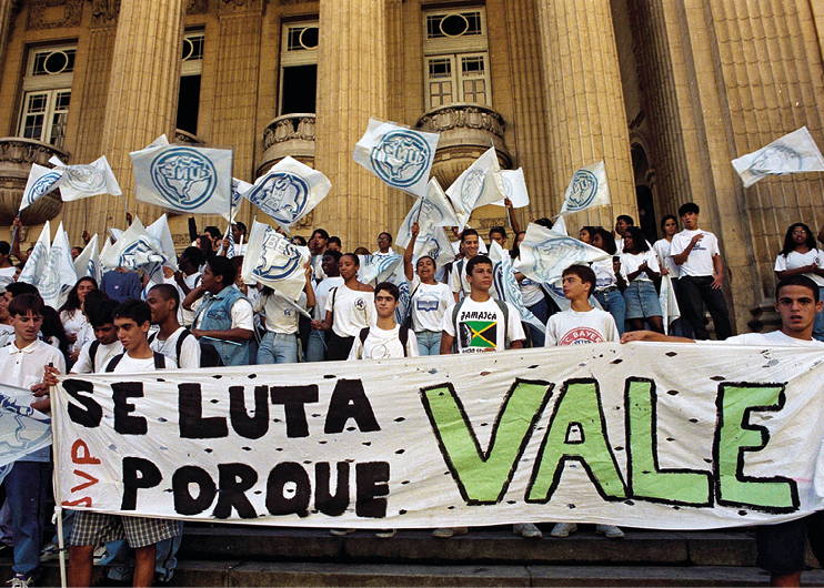 Fotografia. Em frente a um edifício de cor clara, com colunas e janelas brancas altas com balcão, um grupo de pessoas jovens, meninos e meninas, em uma manifestação. Eles usam camisetas brancas, calças jeans na cor azul clara e acenam bandeiras na cor branca.  À frente do grupo, uma faixa com o texto: 'Se luta porque vale'.