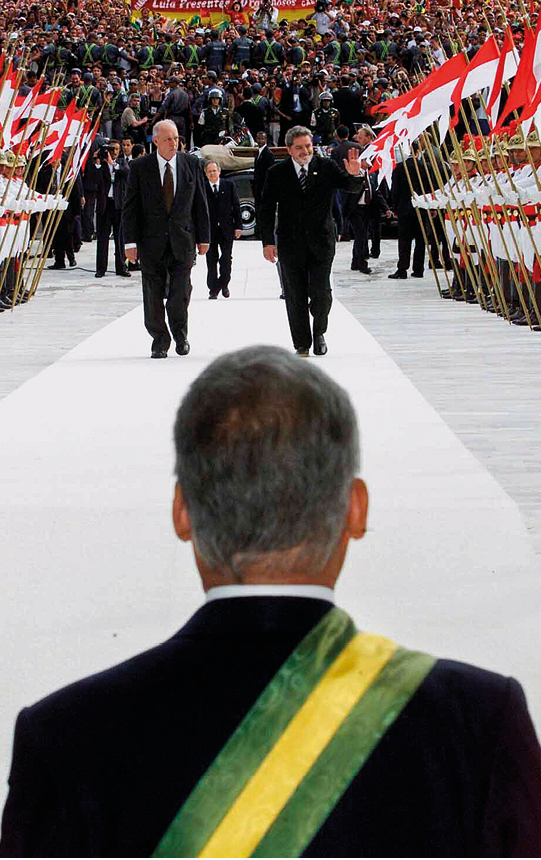 Fotografia. Na parte inferior da imagem, um homem grisalho de cabelos curtos visto de costas. Ele  veste um terno de cor escura e camisa branca e há uma faixa com as cores verde (nas bordas) e amarela (no centro) cruzando suas costas. Diante dele, no centro de uma rampa de cor cinza coberta por um tapete branco há dois homens em pé.
À esquerda, um homem calvo, com cabelos ralos, curtos e brancos, que veste um terno escuro, camisa branca e gravata escura. À direita, um homem de cabelos cheios, curtos e grisalhos, barba grisalha no rosto, que veste um terno escuro, camisa branca, gravata listrada e broche na lapela. Ele está sorrindo e acena com uma das mãos. 
Os homens estão cercados por ambos os lados por militares trajando fardas brancas, com faixas vermelhas na cintura, botas pretas de cano alto, capacetes dourados em forma de dragão com uma pena vermelha na ponta. Cada um dos militares segura a haste dourada de uma bandeira com duas faixas horizontais, nas cores vermelha e branca. Ao fundo, há uma multidão.