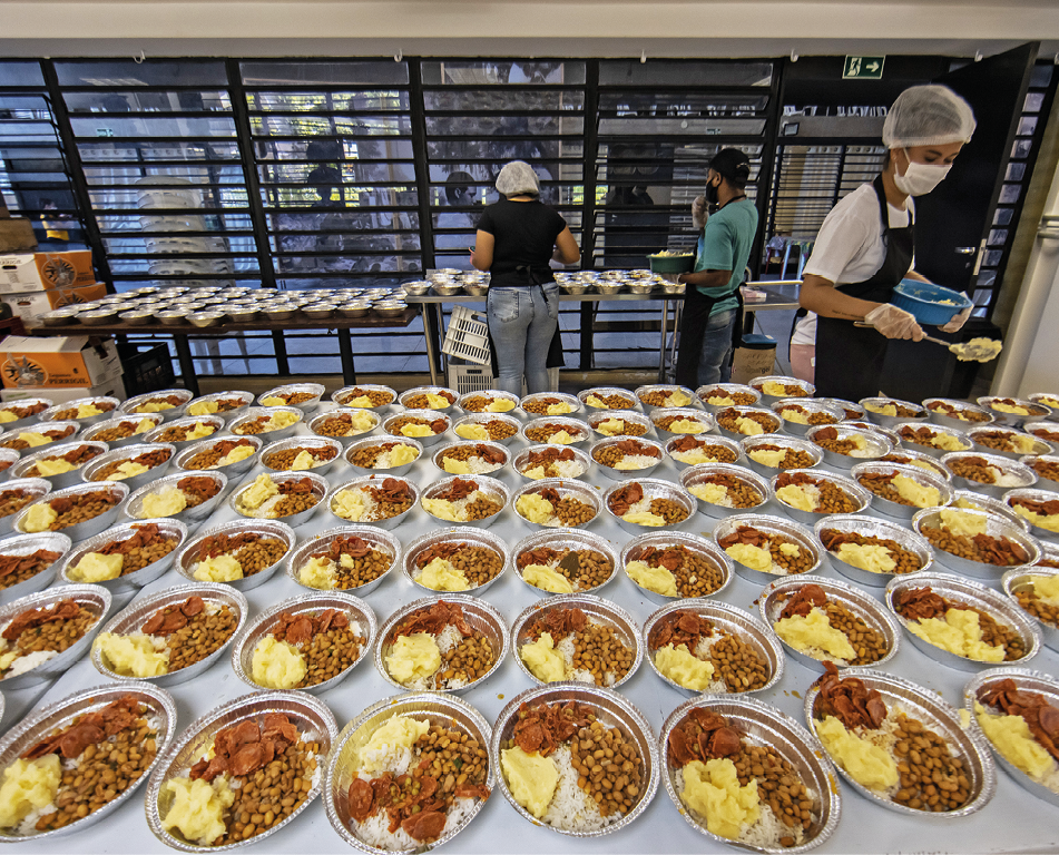 Fotografia. Sobre uma bancada ampla, dezenas de refeições (com arroz, feijão, purê de batatas e carne com molho) dispostas em recipientes redondos e metálicos. 
Ao fundo, à direita, uma jovem mulher com os cabelos presos está em pé, vestindo um avental escuro, camiseta branca, luvas nas mãos, touca branca em sua cabeça e máscara em seu rosto. Ela segura um recipiente azul com alimento preparado em uma das mãos e uma colher na outra mão, montando uma refeição em um recipiente arredondado e metálico.
Ao fundo, no centro da imagem, vistos de costas, uma mulher e um homem vestindo  aventais escuros dispõem alimentos preparados em recipientes metálicos dispostos em uma grande bancada de metal. No canto esquerdo, caixas de vegetais.