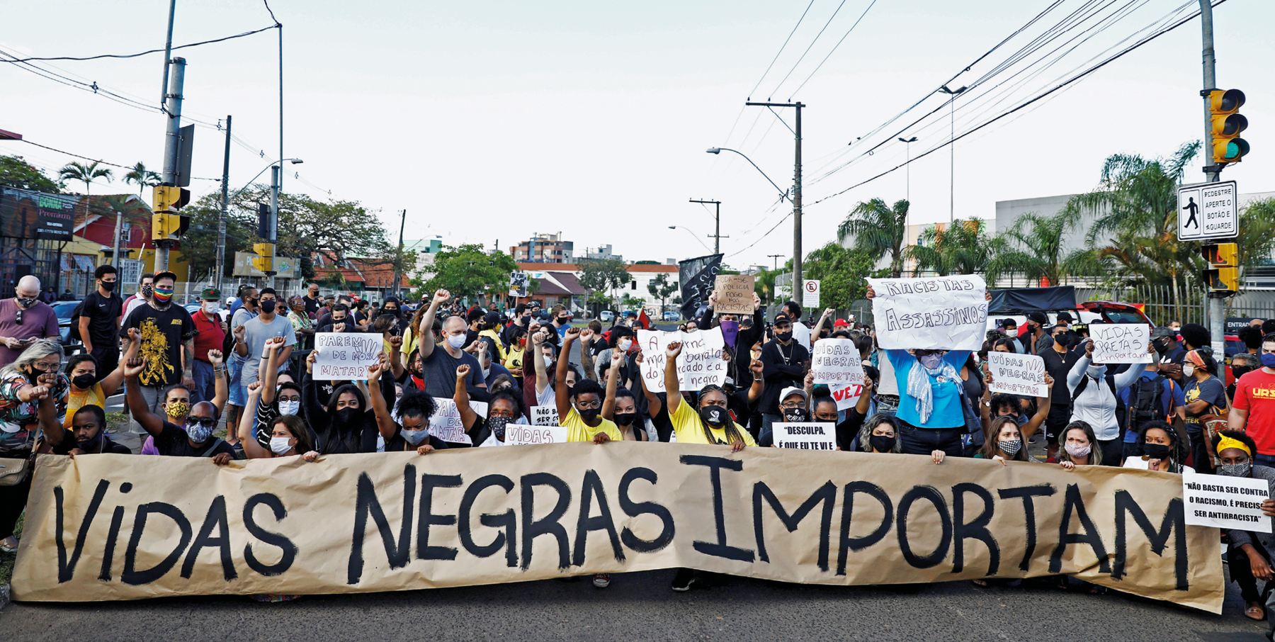 Fotografia. Em uma via pública, uma multidão de pessoas em manifestação, portando cartazes e faixas, com destaque para o texto: 'Vidas negras importam'.