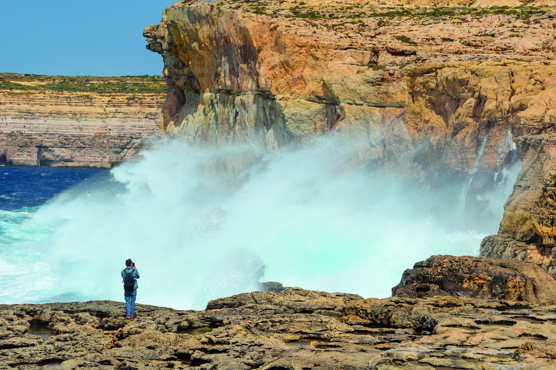 Fotografia. Vista de uma grande onda batendo em uma formação rochosa que fica de frente para o mar. Sobre as rochas, uma pessoa observa a onda.