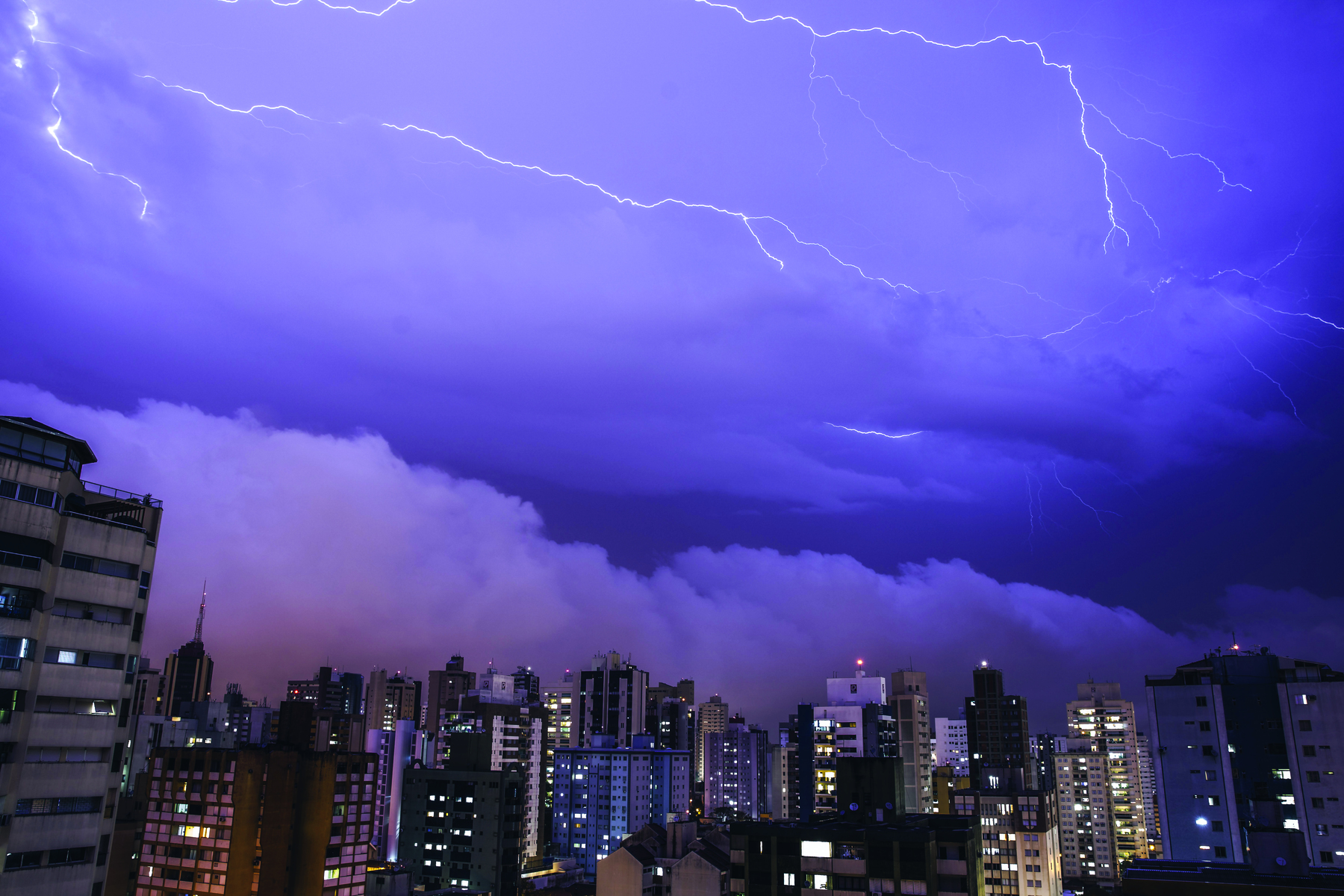 Fotografia. Área com muitos prédios altos, com luzes acesas nas janelas. O céu está nublado, escuro, com  nuvens e raios.