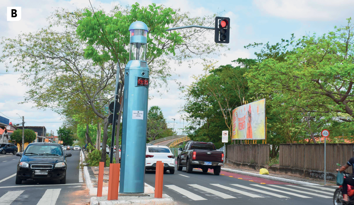 Fotografia B. Vista de uma rua asfaltada, com faixa de pedestres, semáforo, árvores no canteiro central e carros nas ruas.