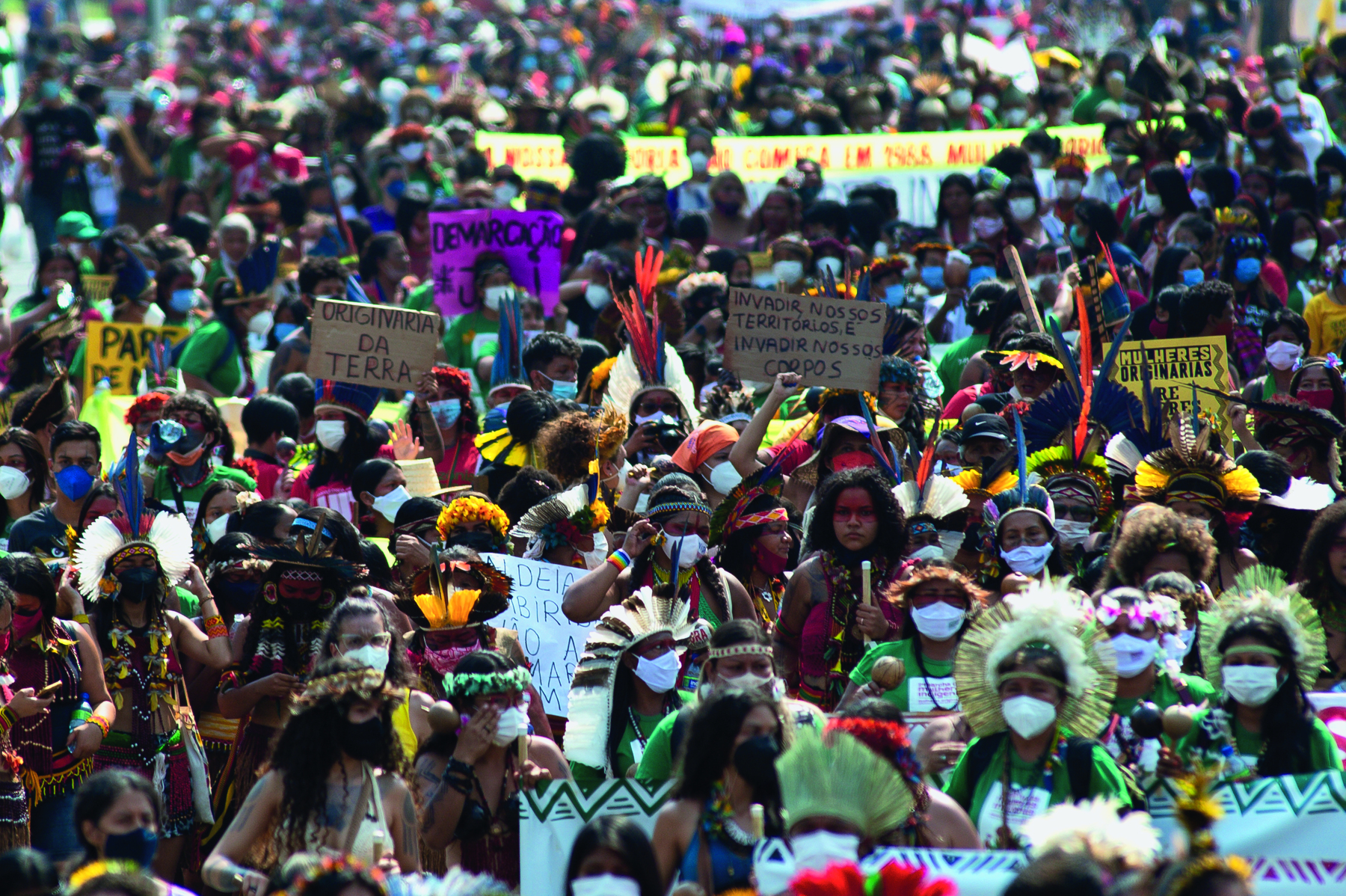 Fotografia. Multidão de pessoas aglomeradas, entre elas, muitas mulheres. As pessoas usam máscaras de proteção sobre a boca e o nariz, cocares de penas coloridas na cabeça, pintura no rosto e seguram cartazes com frases.