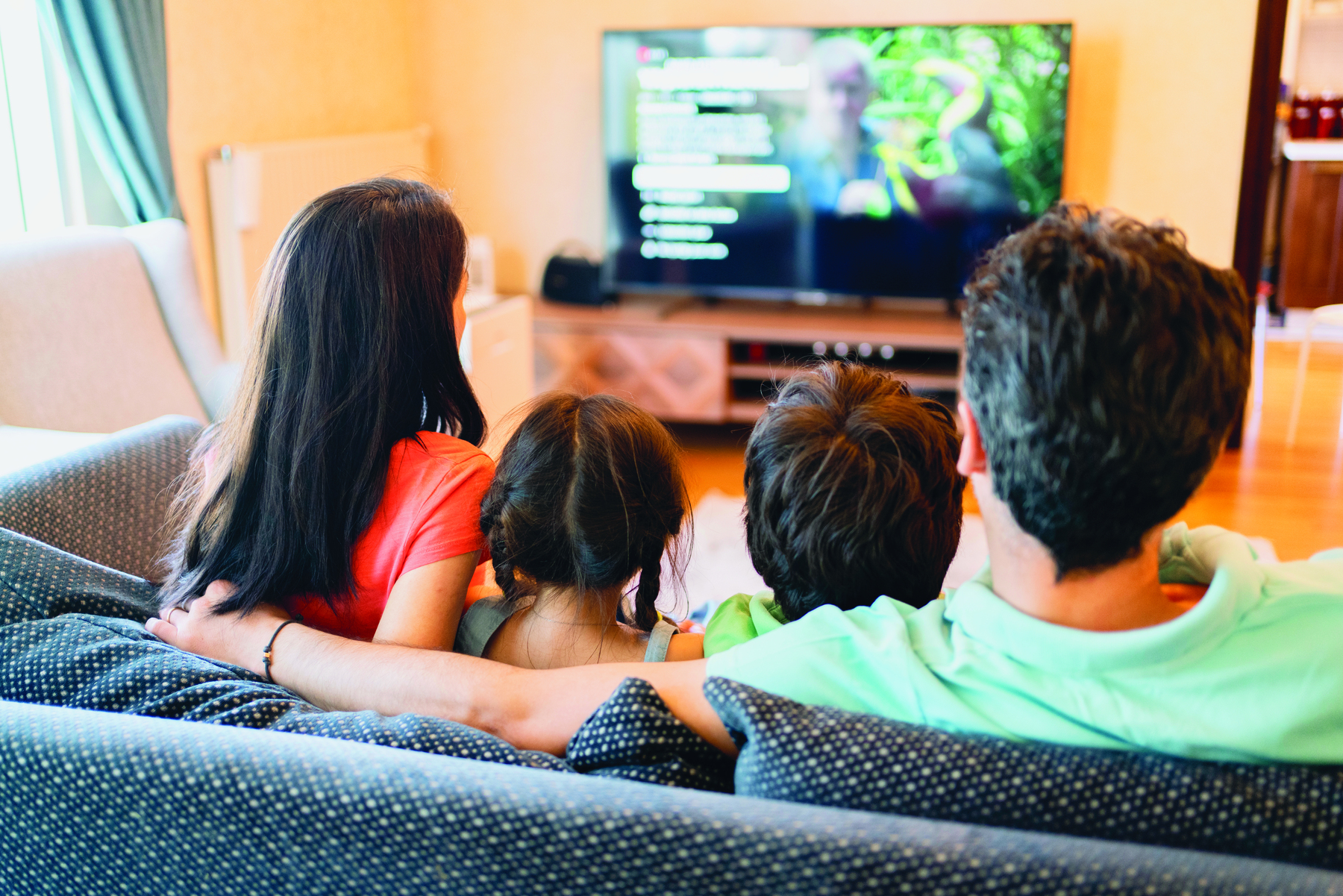 Fotografia. Um casal formado por um homem e uma mulher e duas crianças sentados no sofá. Eles olham para a televisão que está a frente deles.